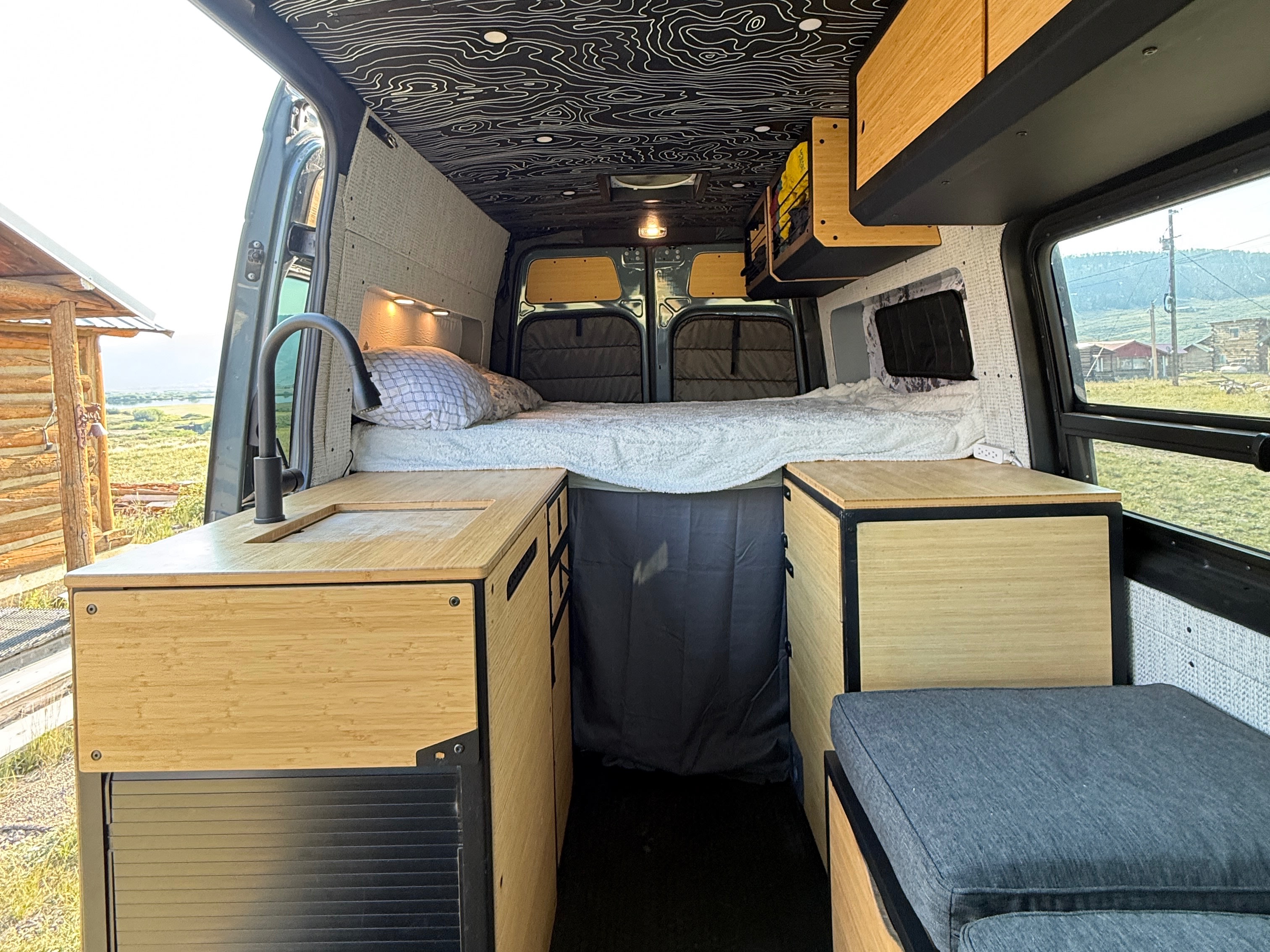 Modern rv rental van conversion with bamboo kitchen, comfy bed, and map ceiling, offering scenic views outside.. Mercedes-Benz Sprinter 2021