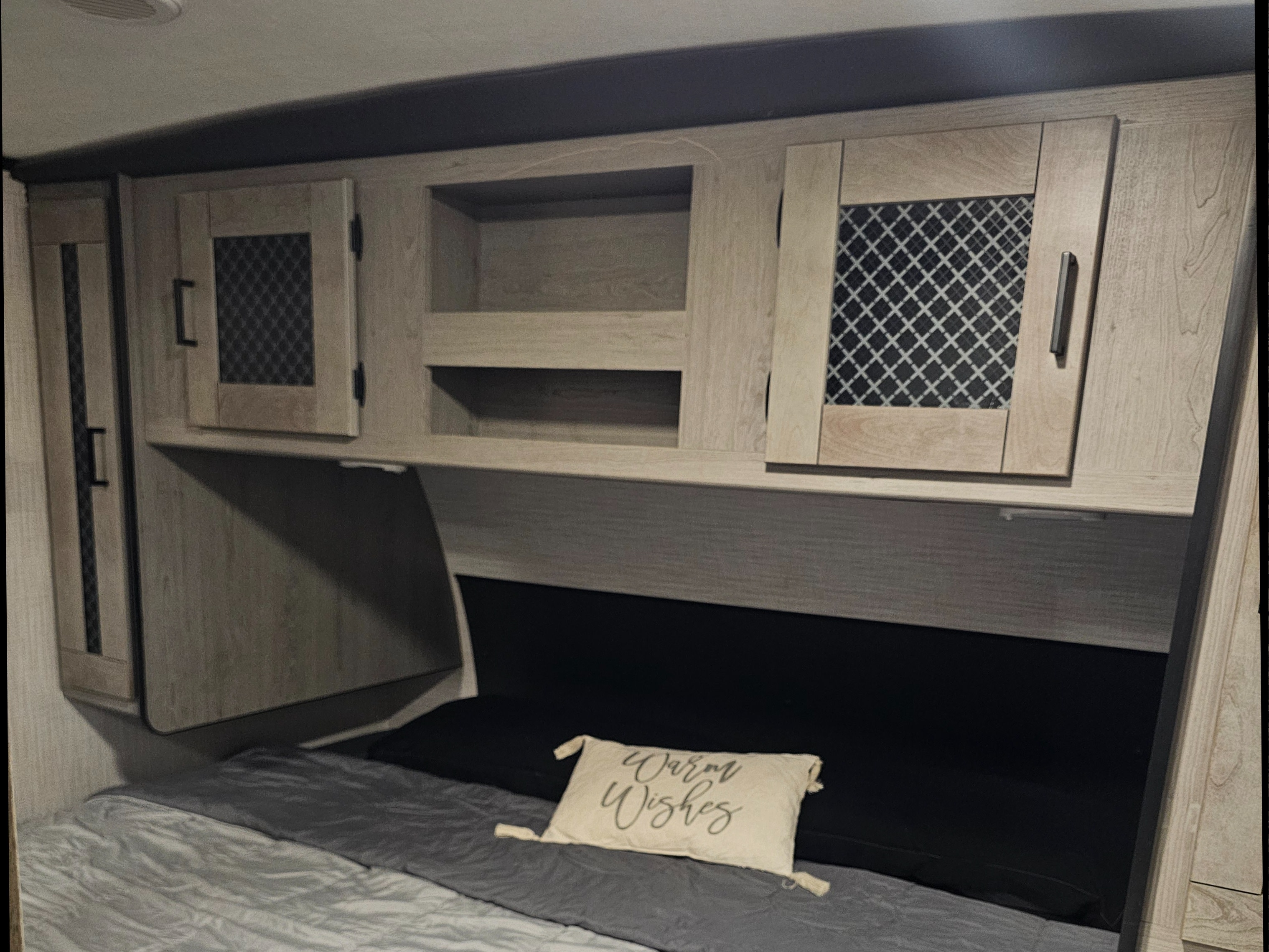 RV rental bedroom with light wood overhead cabinets (some with mesh), open cubby, and bed with gray bedding and a 'Warm Wishes' pillow.. Heartland RVs Mallard 2021