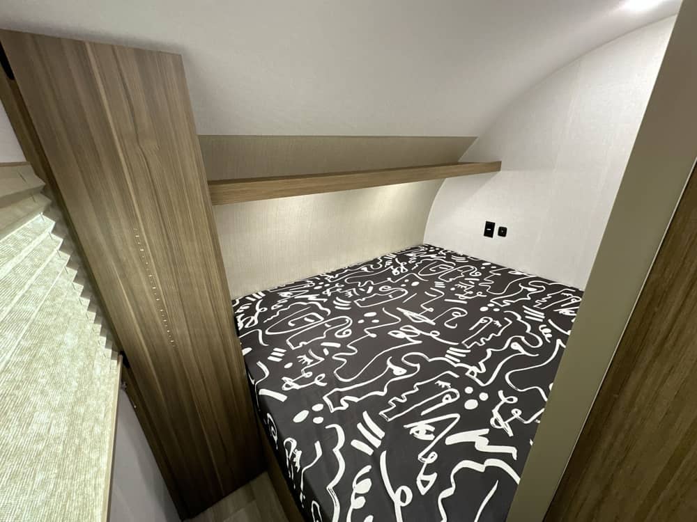 Interior of an RV rental bedroom showing a bed with black and white linen, a storage shelf, and wall outlets.. Forest River Wildwood FSX 2026