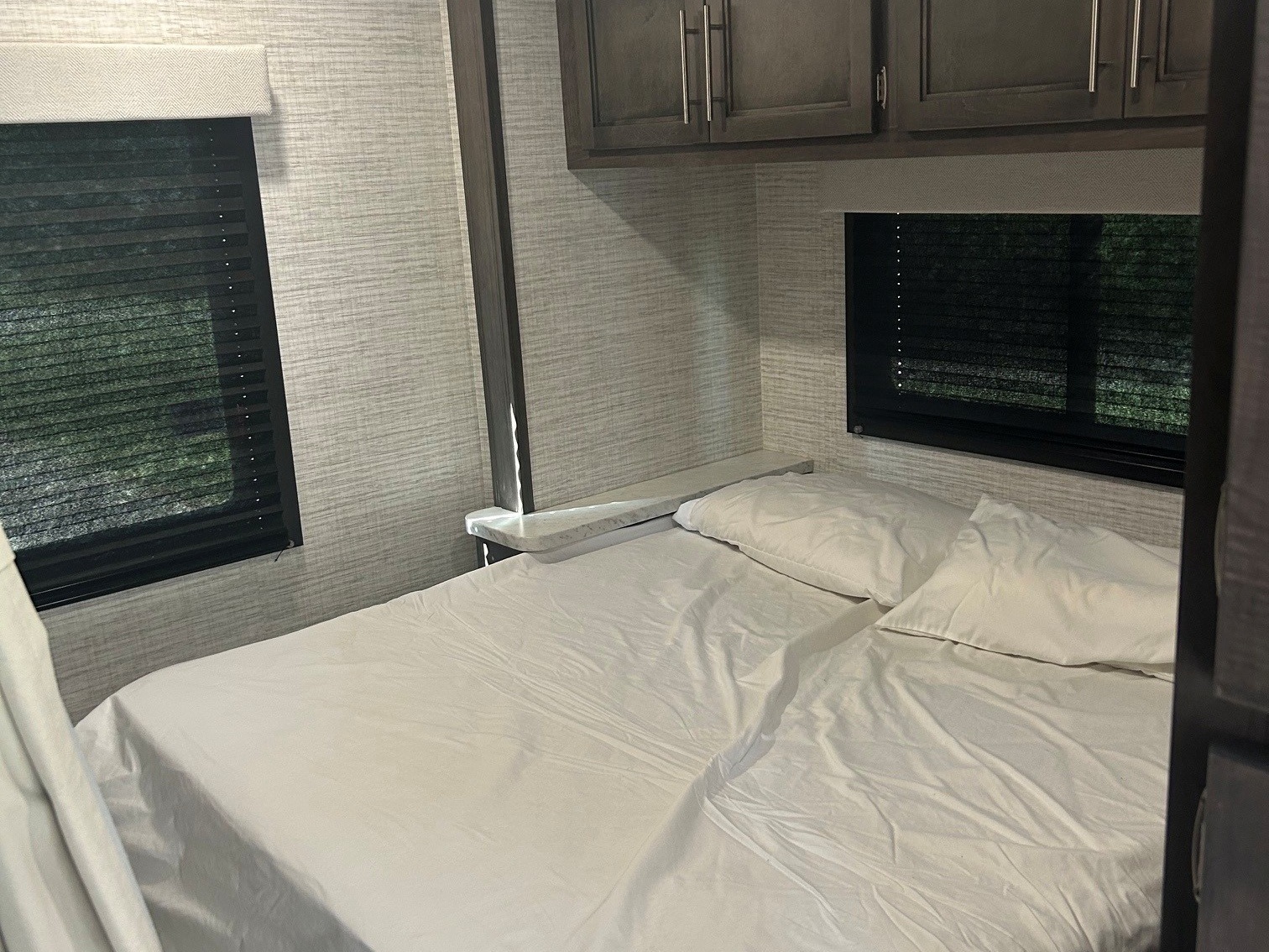 Cozy rv rental bedroom with fresh white linens, dark blinds on windows, and overhead cabinets. Ideal for travel comfort.. Jayco Redhawk SE 2025
