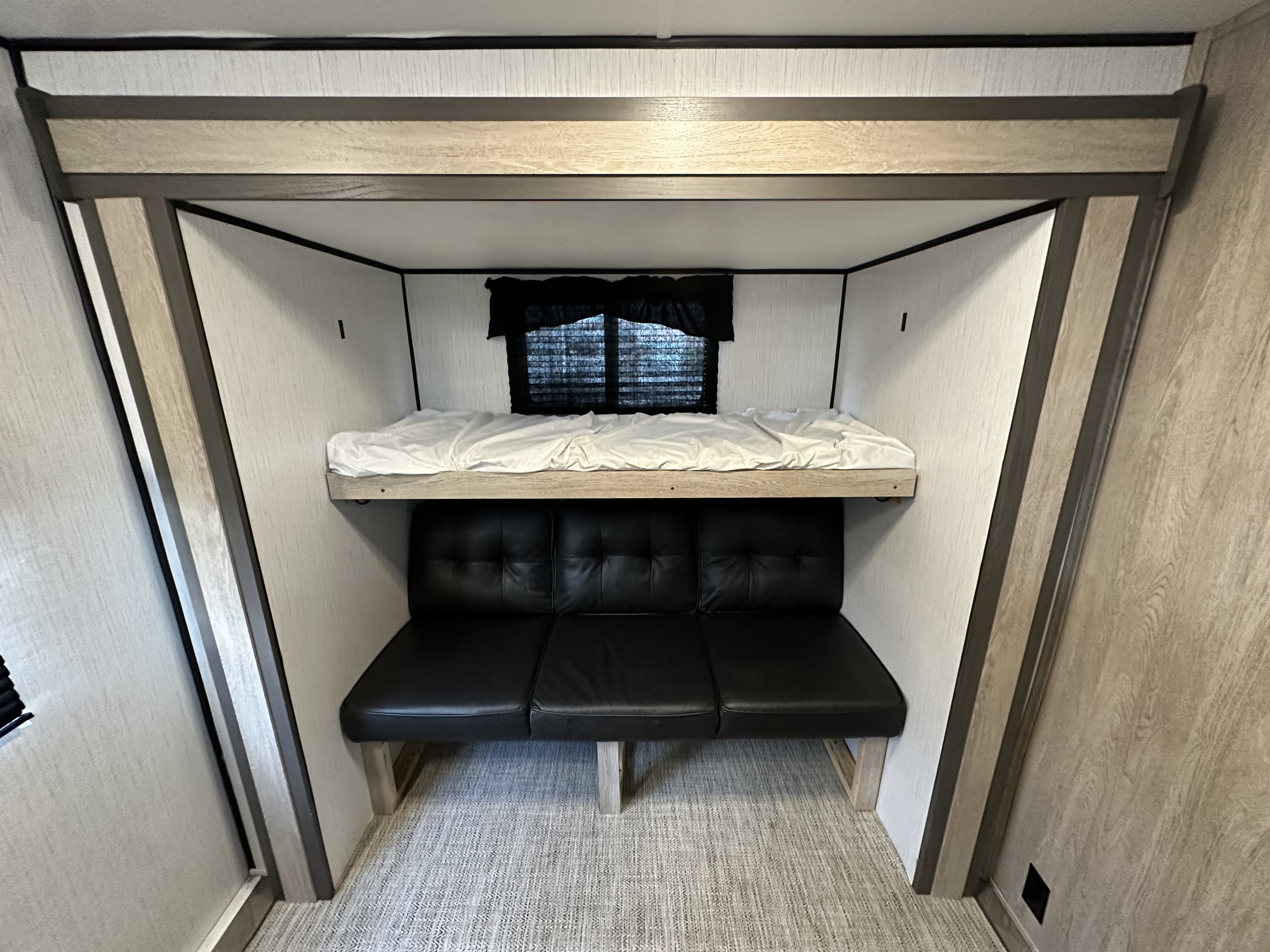 Inside an RV rental: a top bunk bed with white sheets sits above a black faux leather futon. Light walls and wood trim.. Heartland Prowler 2022