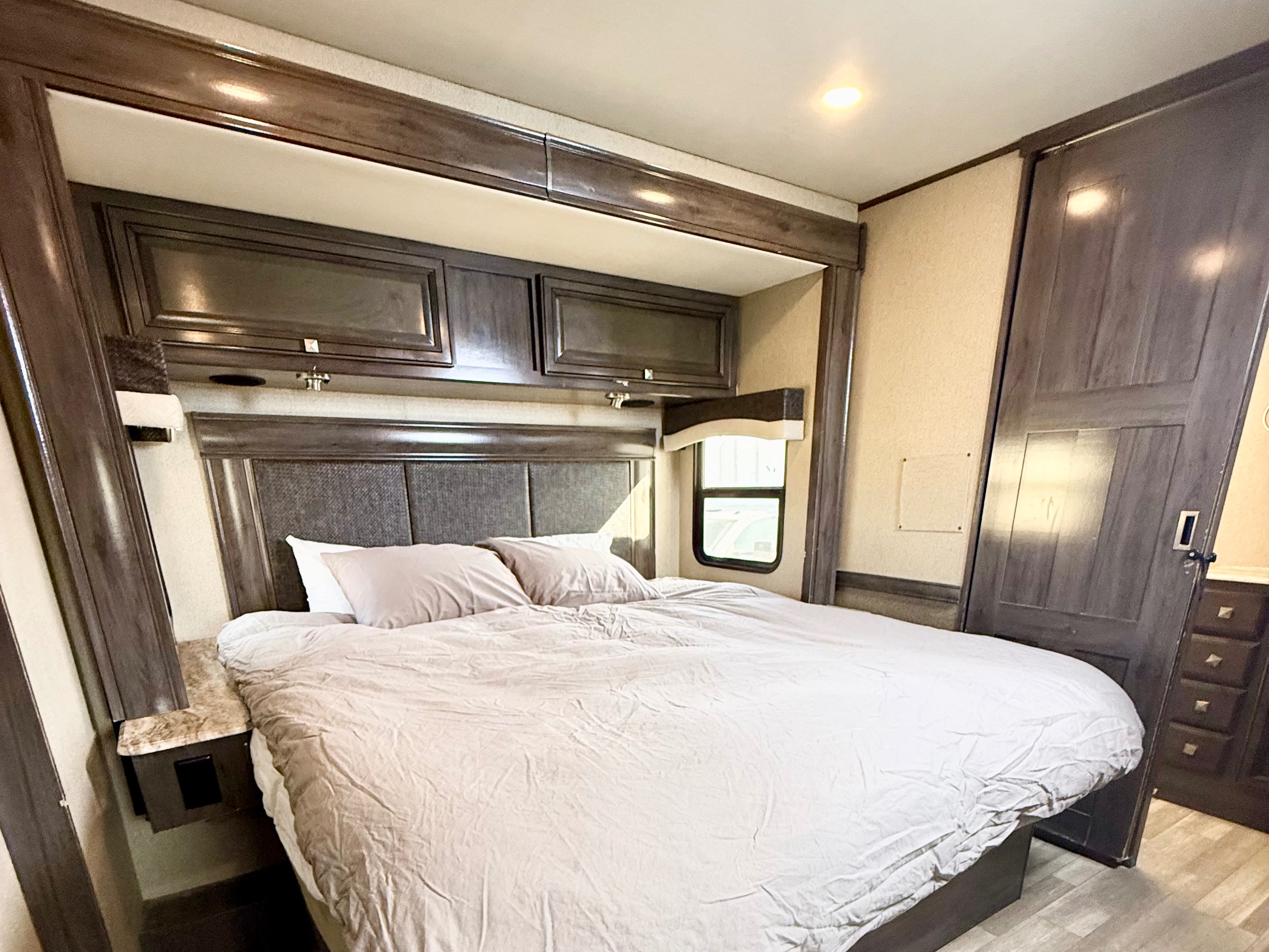 RV rental bedroom: large bed, dark wood headboard, overhead cabinets, and window. A cozy, well-lit interior for travel.. Flair-Fleetwood Flair-Fleetwood Motorhome 2020