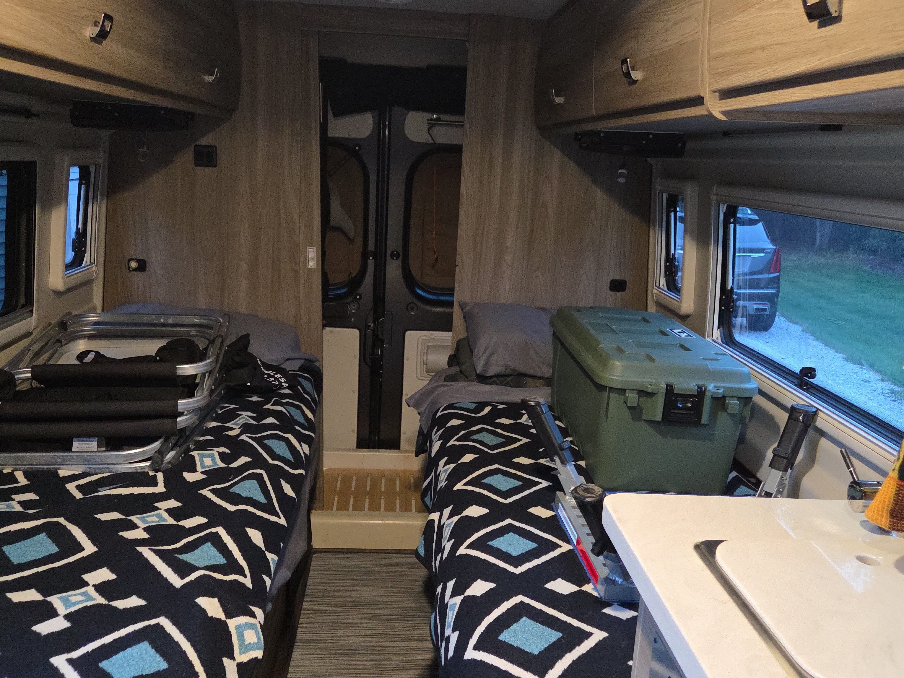 RV rental interior shows twin beds with patterned blankets, a green storage box, and a white kitchen counter.. Winnebago Travato 2023