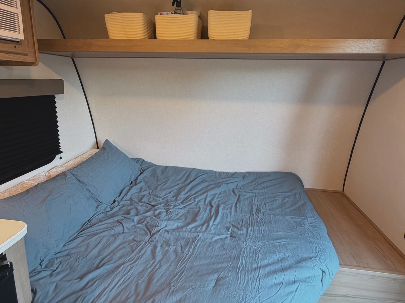 Cozy bed in an RV rental, featuring blue bedding, a wooden shelf with woven baskets, and a window with a dark blind.. Keystone RV Coleman 2026