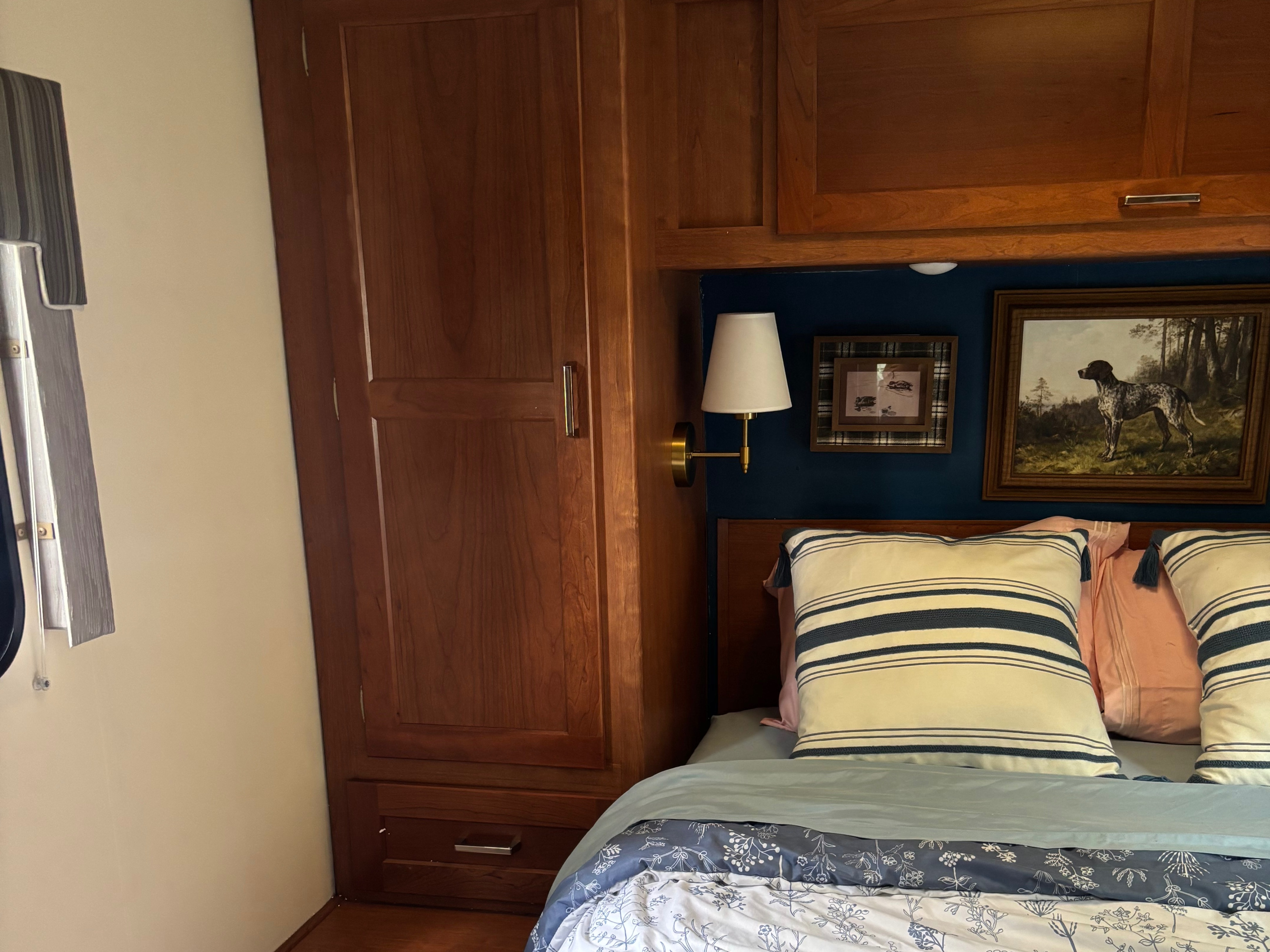 Cozy RV rental bedroom with wooden storage, a soft bed, sconce light, and framed art above the dark blue headboard.. Winnebago Minnie Winnie 2019