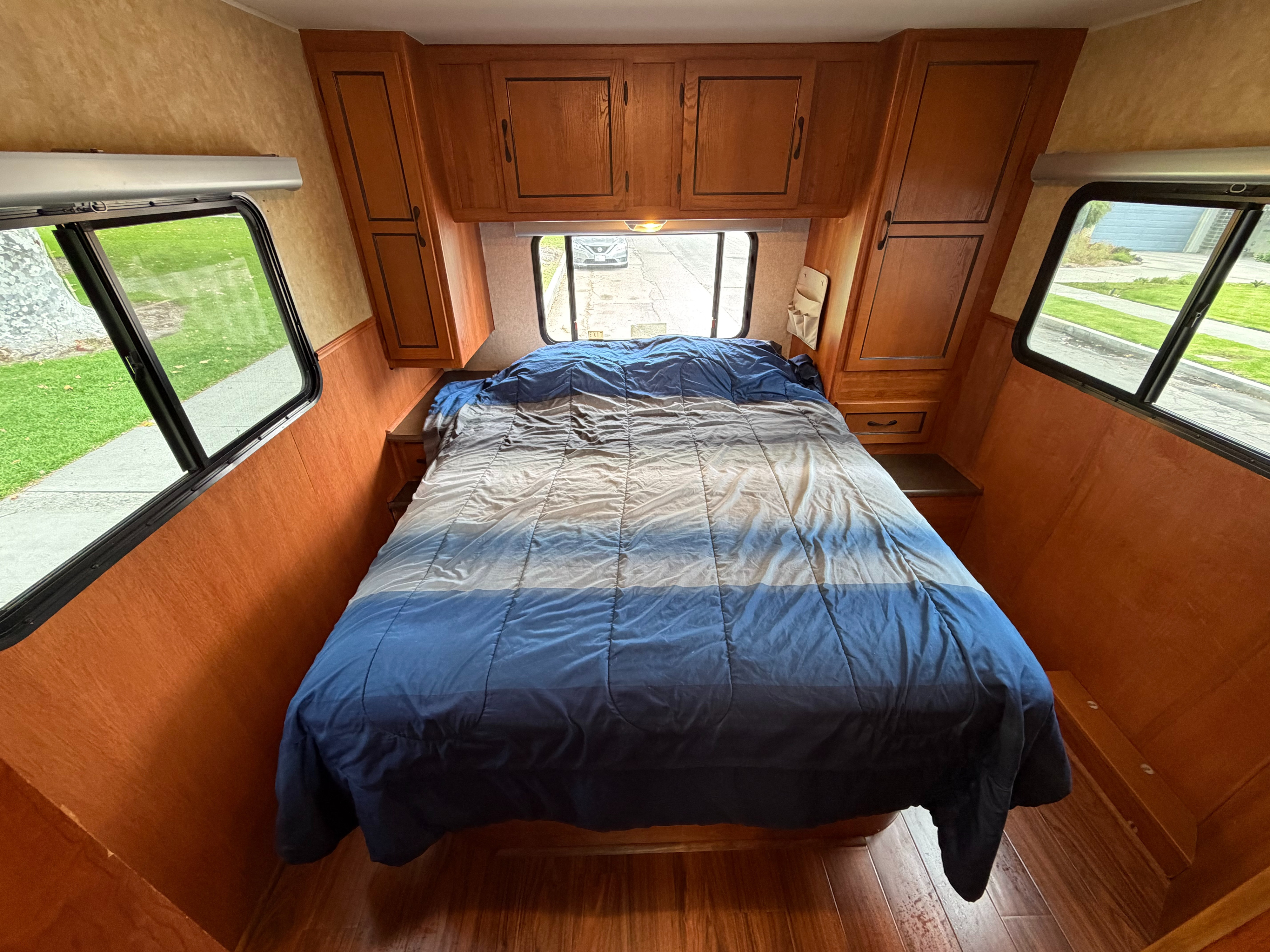 Cozy RV rental bedroom features a blue and gray striped bed, wooden cabinets, and large windows on both sides.. Coachmen Freelander 2010
