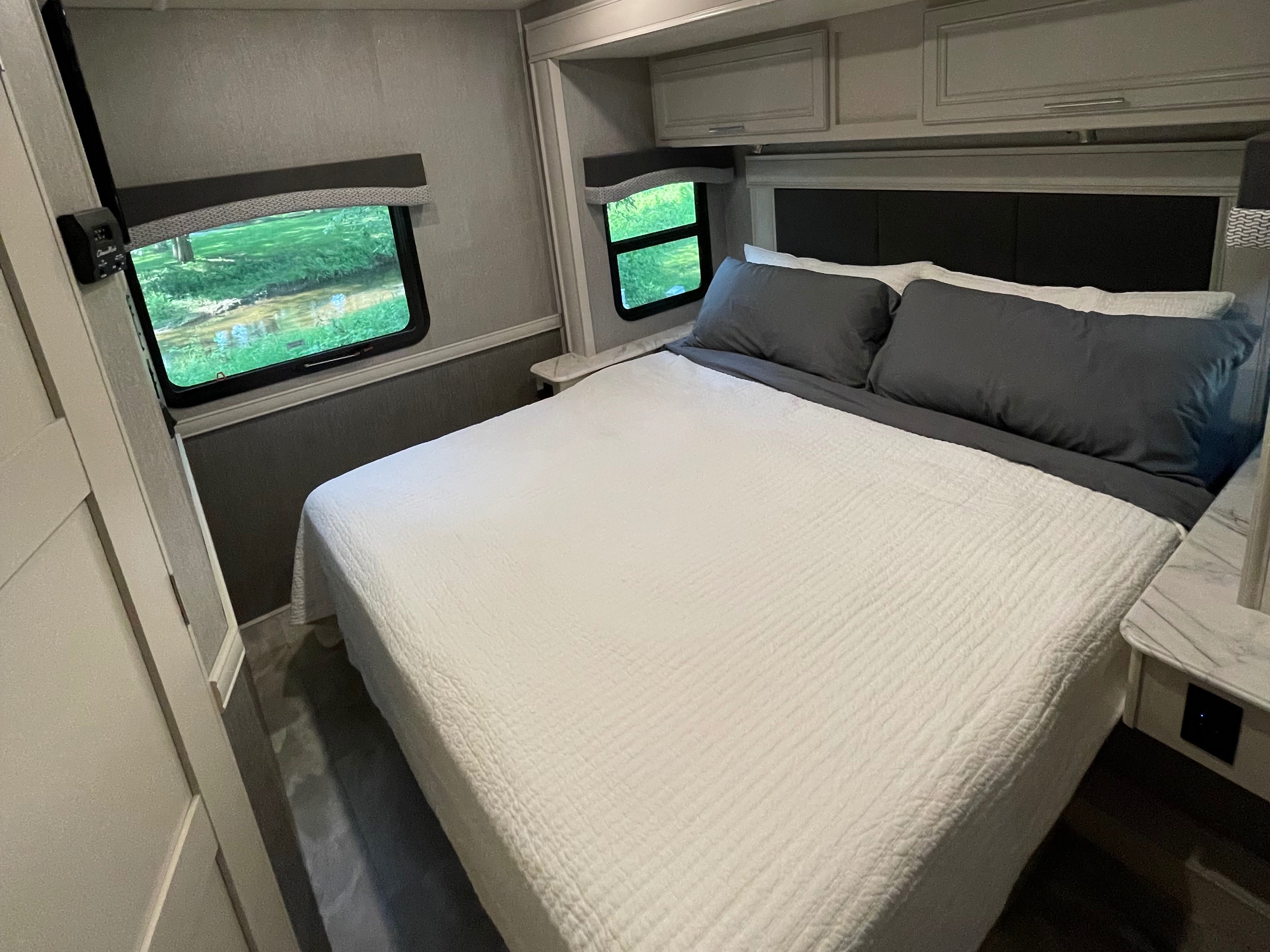 RV rental bedroom: white bed, grey pillows, and windows overlooking lush greenery. Features modern cabinets and nightstands.. Flair-Fleetwood Flair-Fleetwood Motorhome 2022