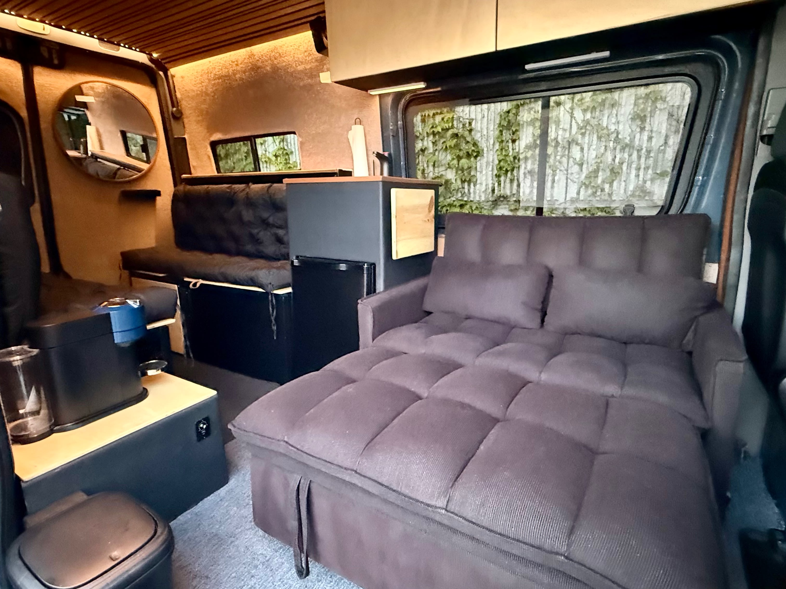 Cozy RV rental interior: extended sofa bed, bench, mini-fridge, and mirror in a warm, wood-accented camper van.. Mercedes-Benz Sprinter 2018