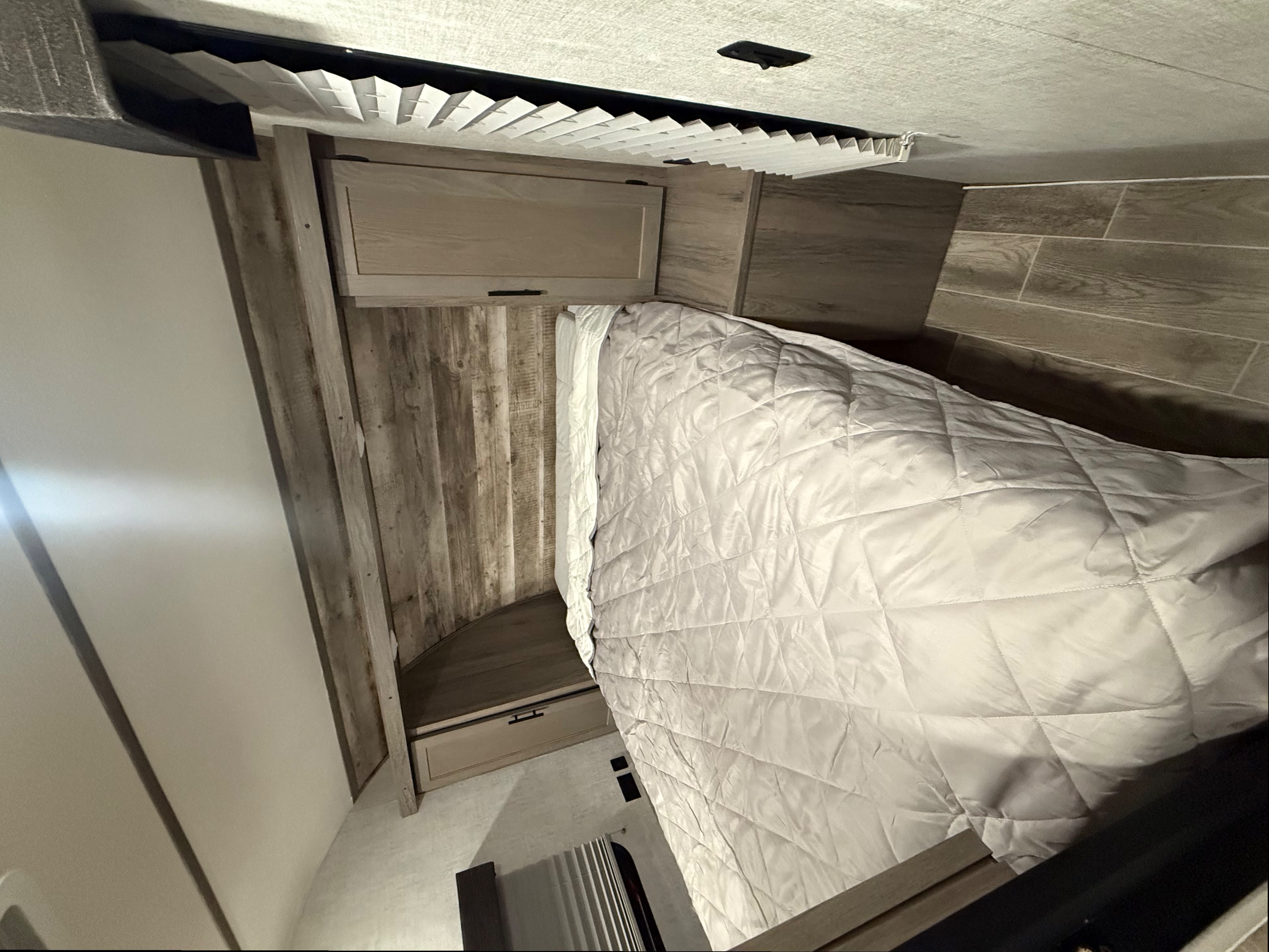 Inside this RV rental bedroom, a neatly made bed with a quilted comforter, wood-paneled walls, and pleated blinds.. Gulf Stream Ameri-Lite 2021