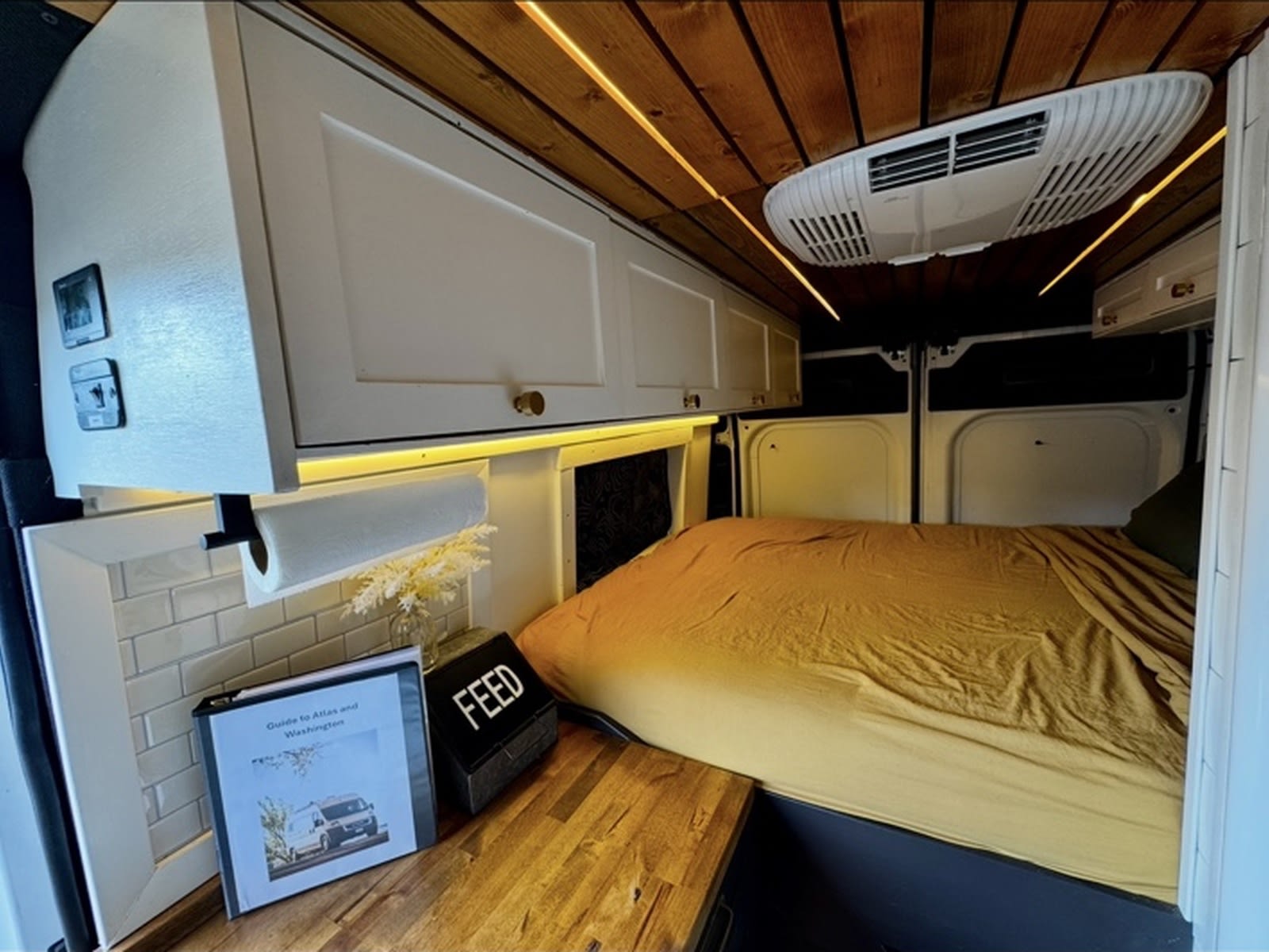 This RV rental showcases a cozy bed, wood-paneled ceiling with LED lights, overhead cabinets, and AC, plus a guide for guests.. Dodge Ram Promaster 2500 Highroof 2020