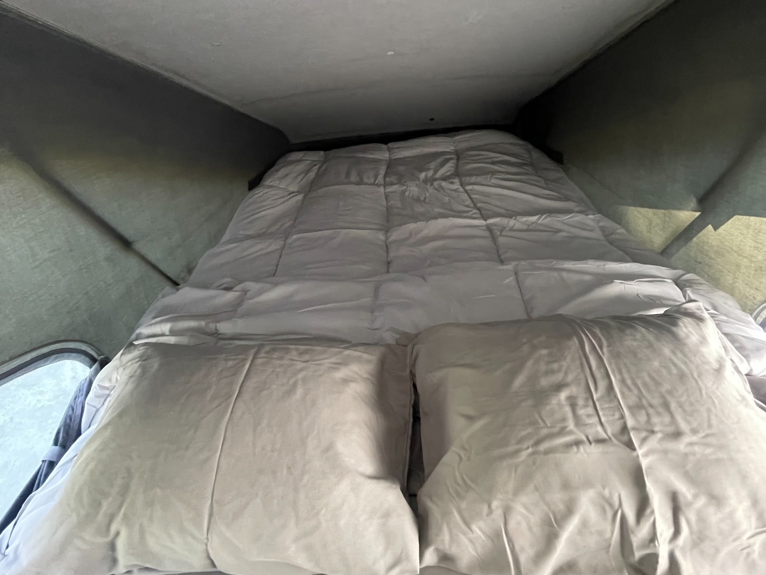 Comfortable bed inside an RV rental's pop-top sleeping area, featuring light gray bedding, pillows, and a comforter.. Toyota Hiace Campervan 1994