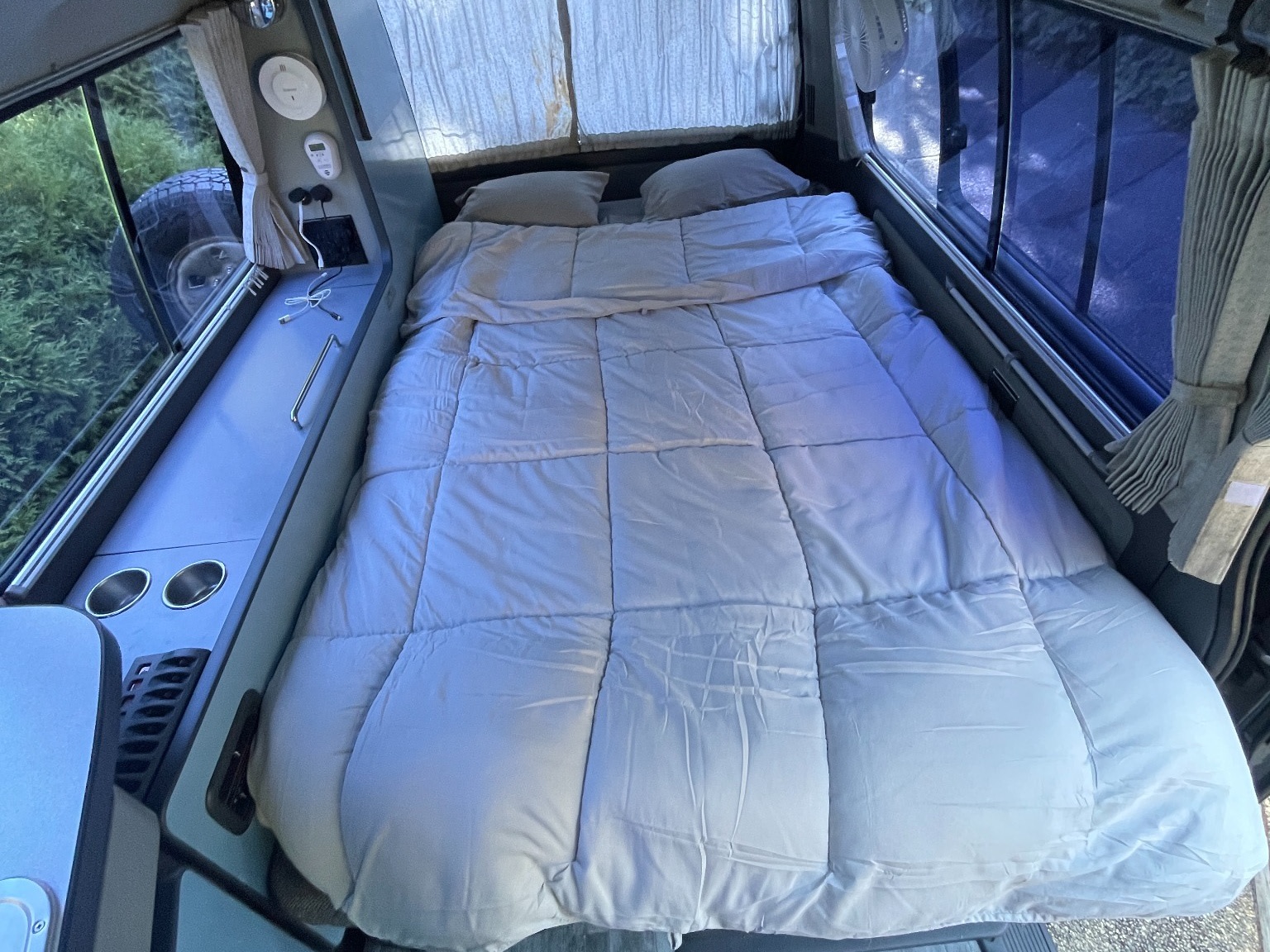 Cozy RV rental's interior: a comfortable bed, side console with charging ports, cup holders, and a view outside.. Toyota Hiace Campervan 1994
