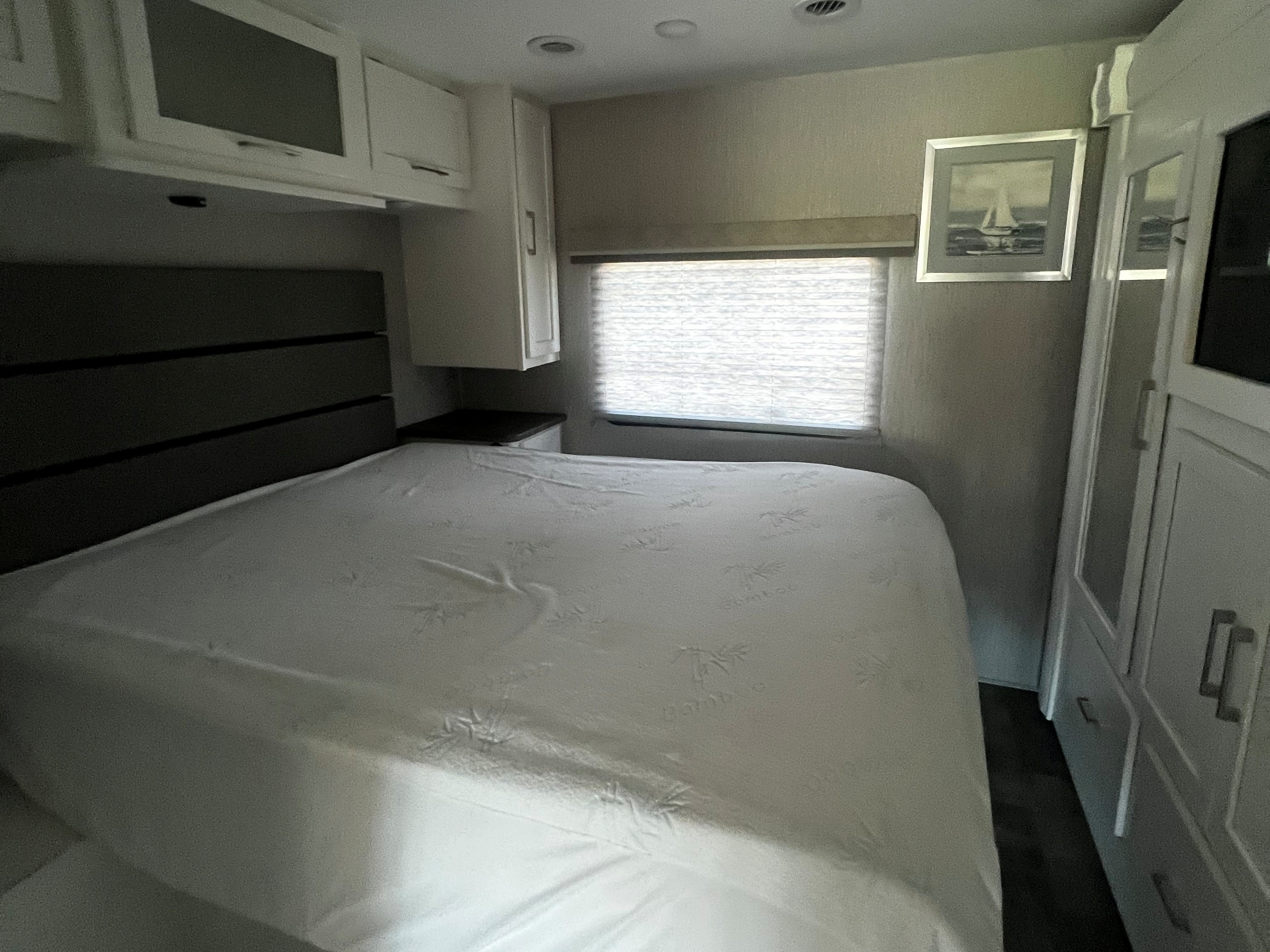 Cozy RV rental bedroom featuring a bed, headboard, overhead cabinets, window with blinds, and sailboat art.. Forest River Coachmen Pursuit 2022