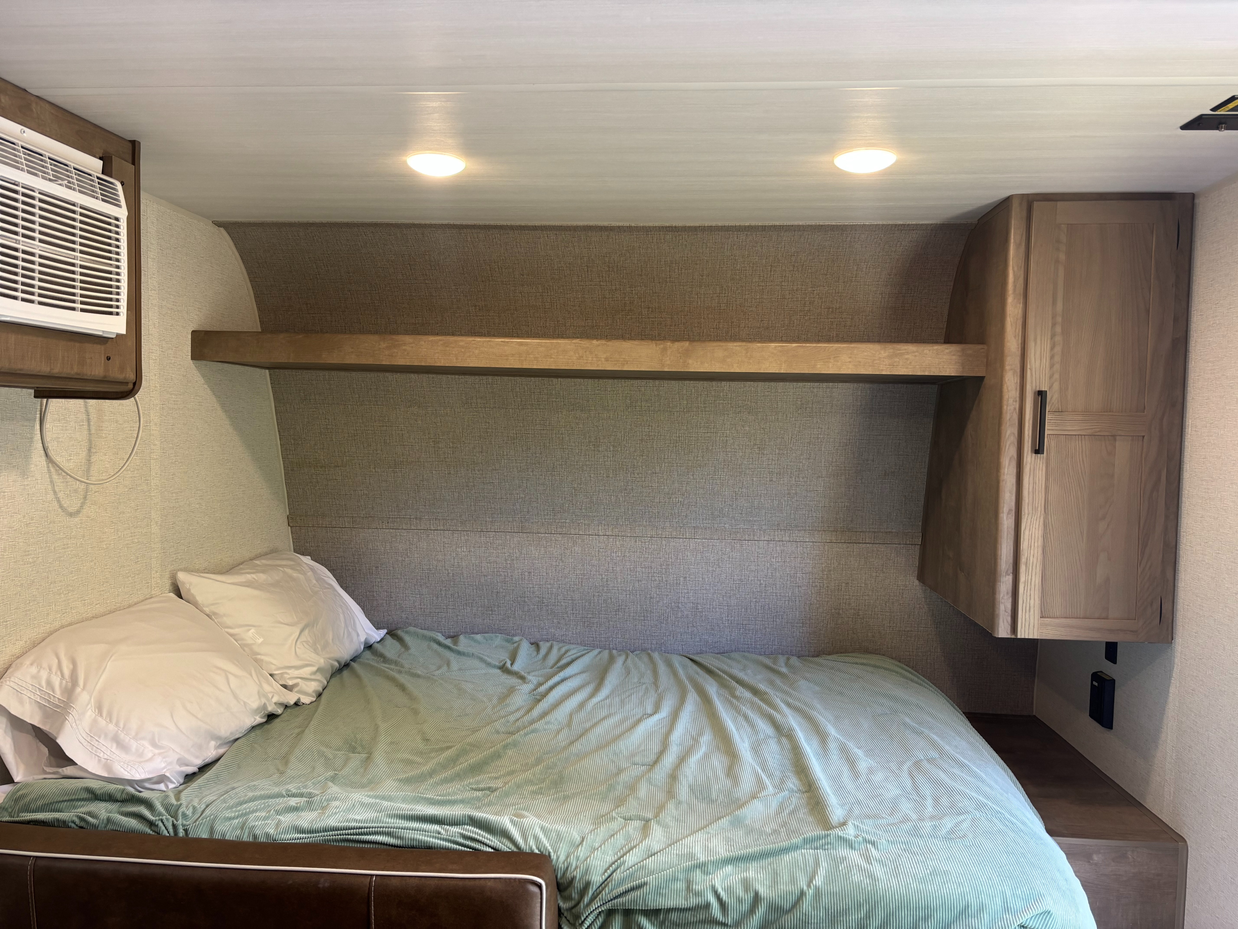 Comfortable RV rental bedroom with teal blanket, overhead lights, AC, and wooden storage.. Keystone RV Springdale 2025