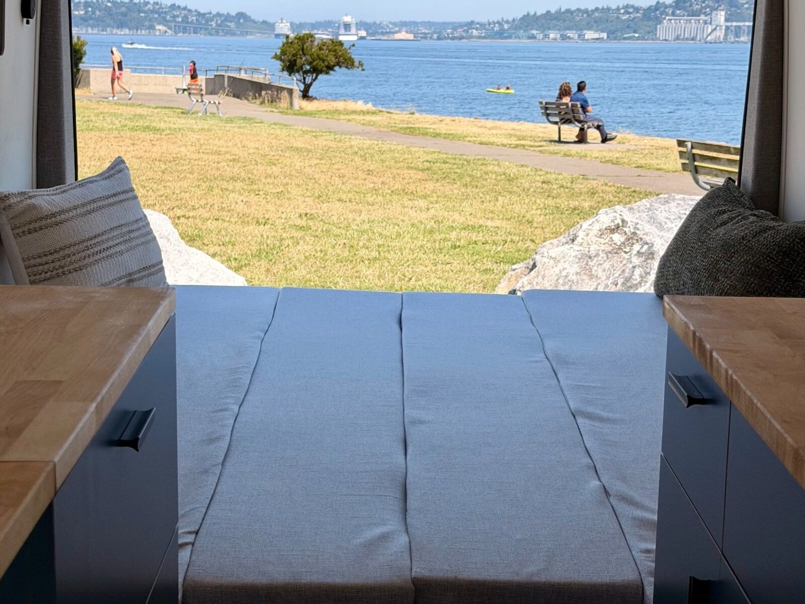 Inside this RV rental, a comfy bed and wood counters overlook a serene waterfront park with people and boats on the water.. FORD TRANSIT T-350 2026