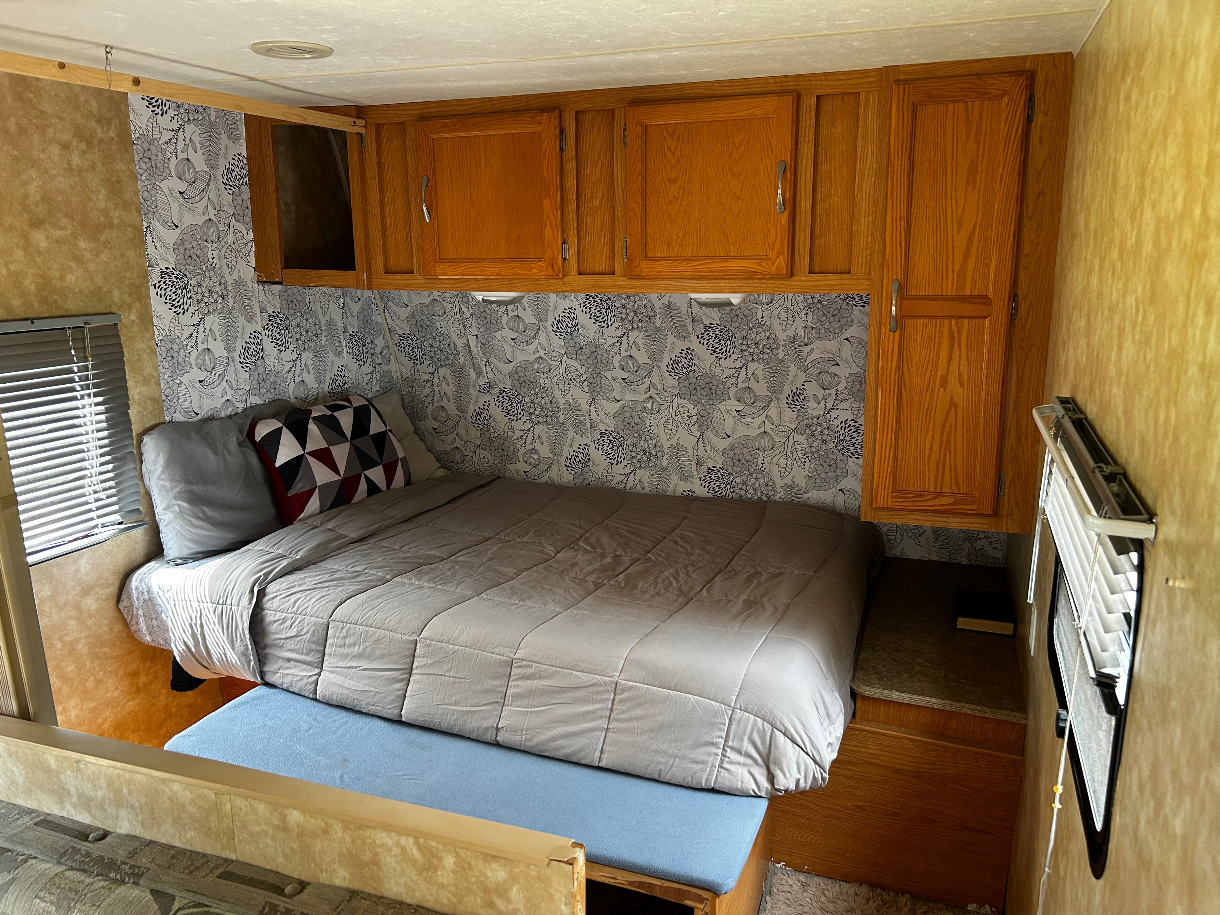 This cozy rv rental bedroom features a bed with gray bedding, floral wallpaper, and wood storage cabinets above. A compact space for travel.. Coachmen Spirit of America 2009