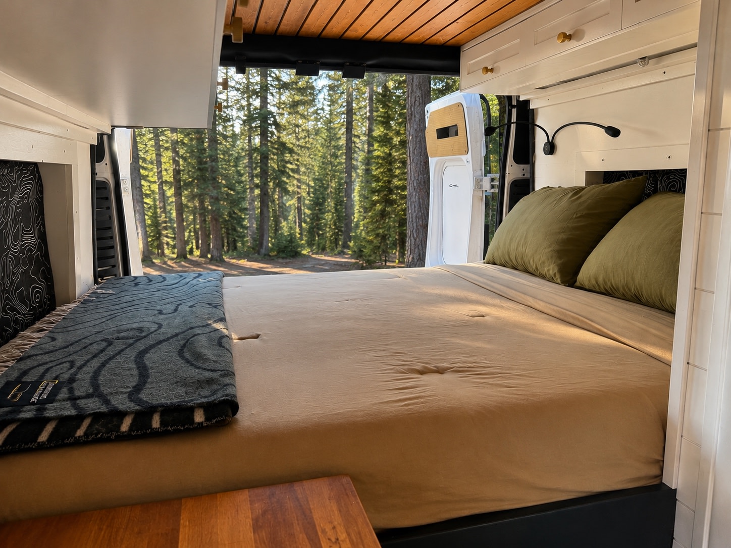 Inside this RV rental bedroom, a comfortable bed offers a stunning view of a sunlit forest through open rear doors.. Dodge Ram Promaster 2500 Highroof 2020