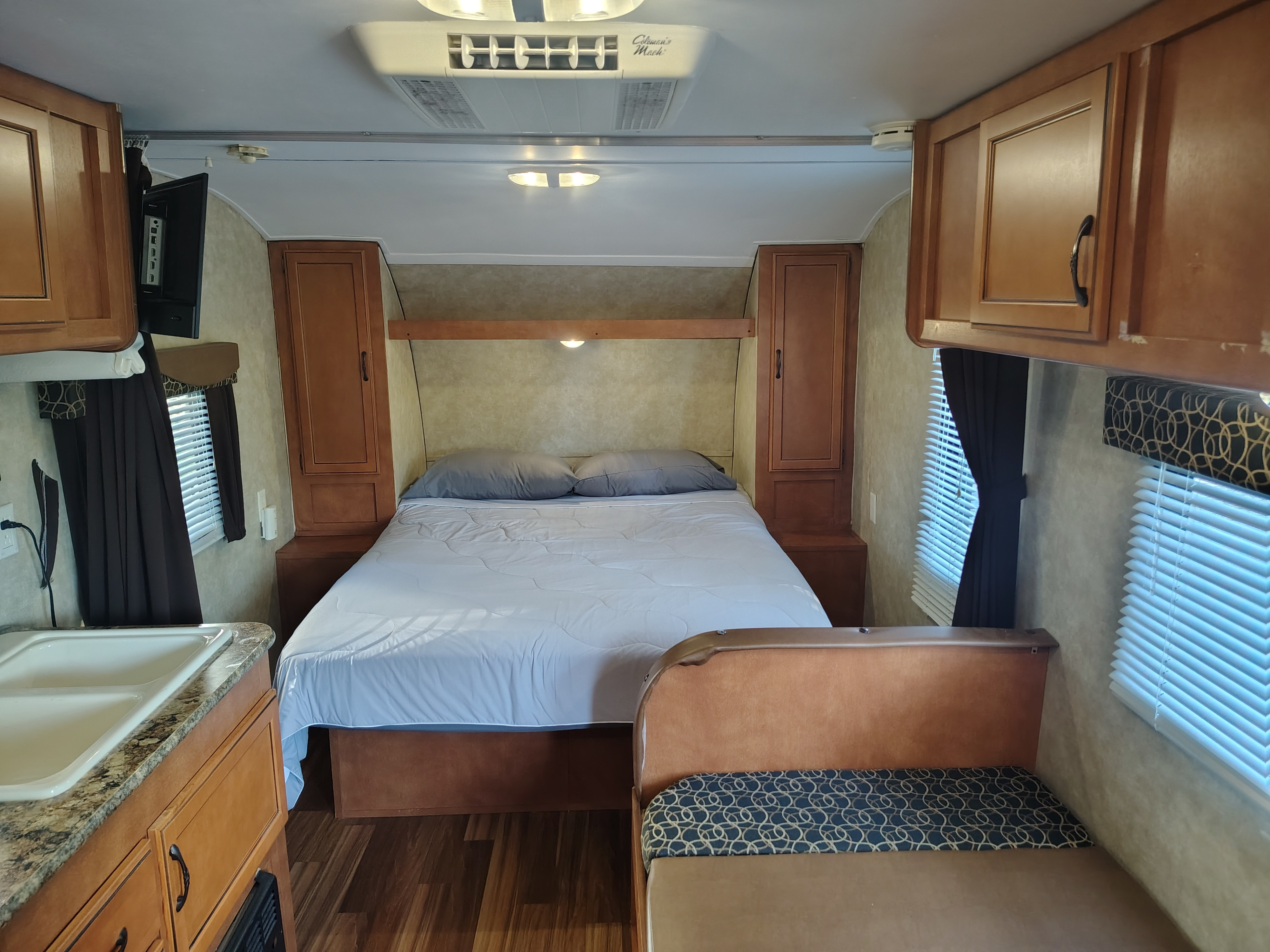 Interior of an RV rental showing a neatly made bed flanked by wardrobes, a kitchen counter with a sink, and a dinette.. Gulf Stream Ameri-Lite 2015