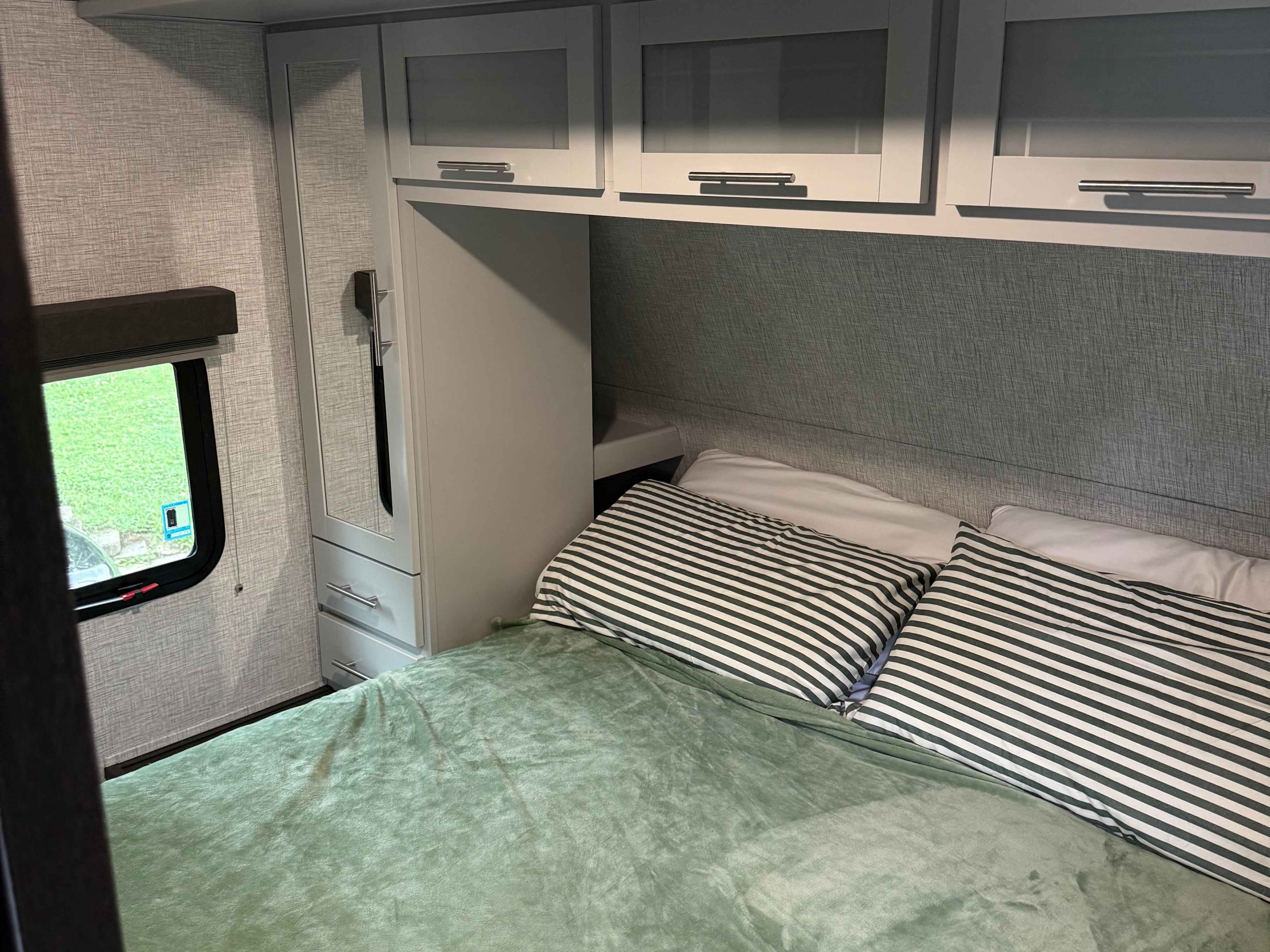 RV rental bedroom featuring a bed with a green blanket, striped pillows, overhead cabinets, and a window view.. Keystone RV Bullet Ultra Lite 2021