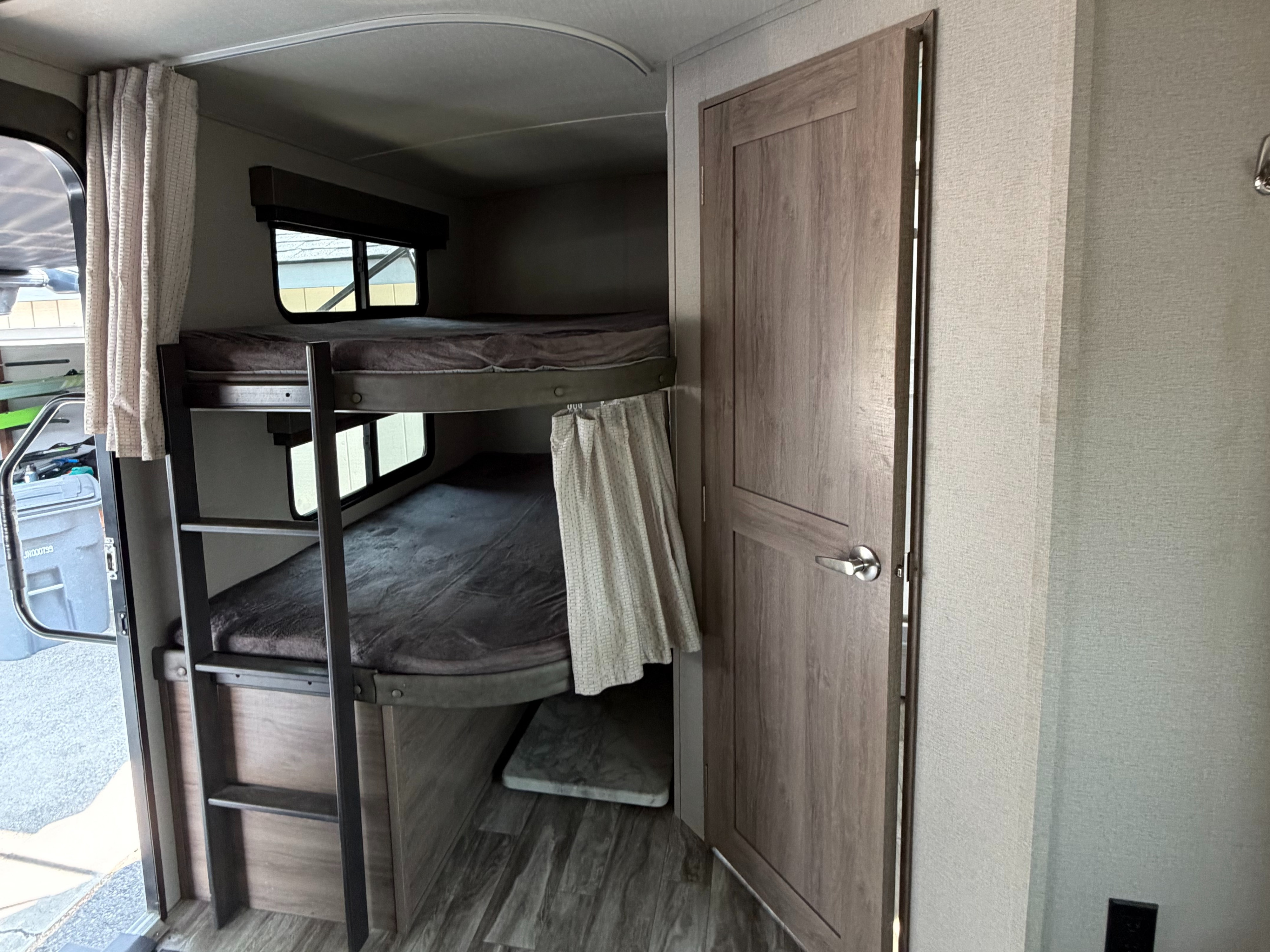 Inside this **rv rental**, a two-tier bunk bed with privacy curtains and windows offers comfortable sleeping. A wood-paneled door is on the right.. Grand Design Imagine 2021