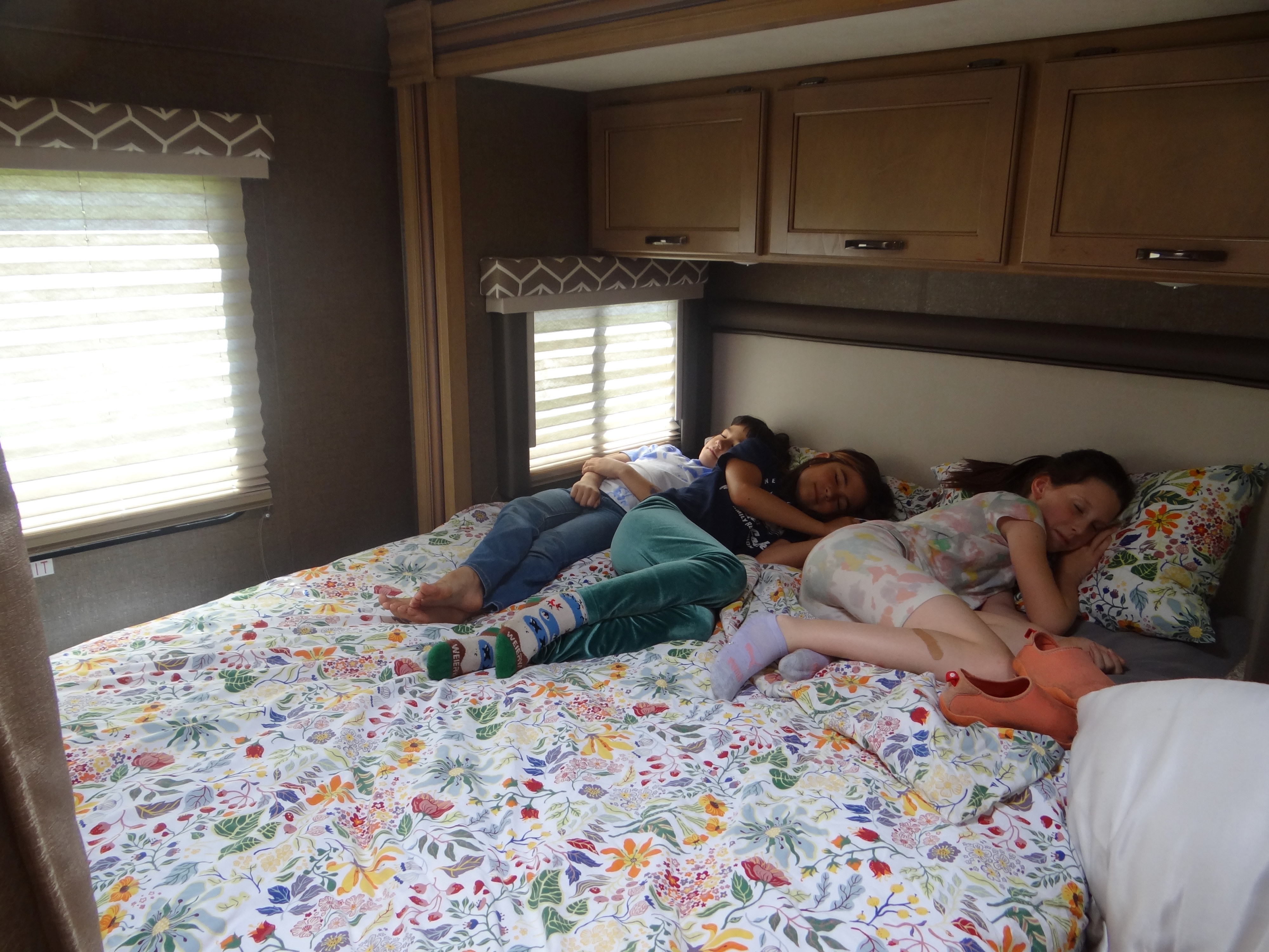 Three children sleeping on a colorful floral bed in an RV rental, with blinds, windows, and overhead cabinets visible.. Thor Daybreak 2019