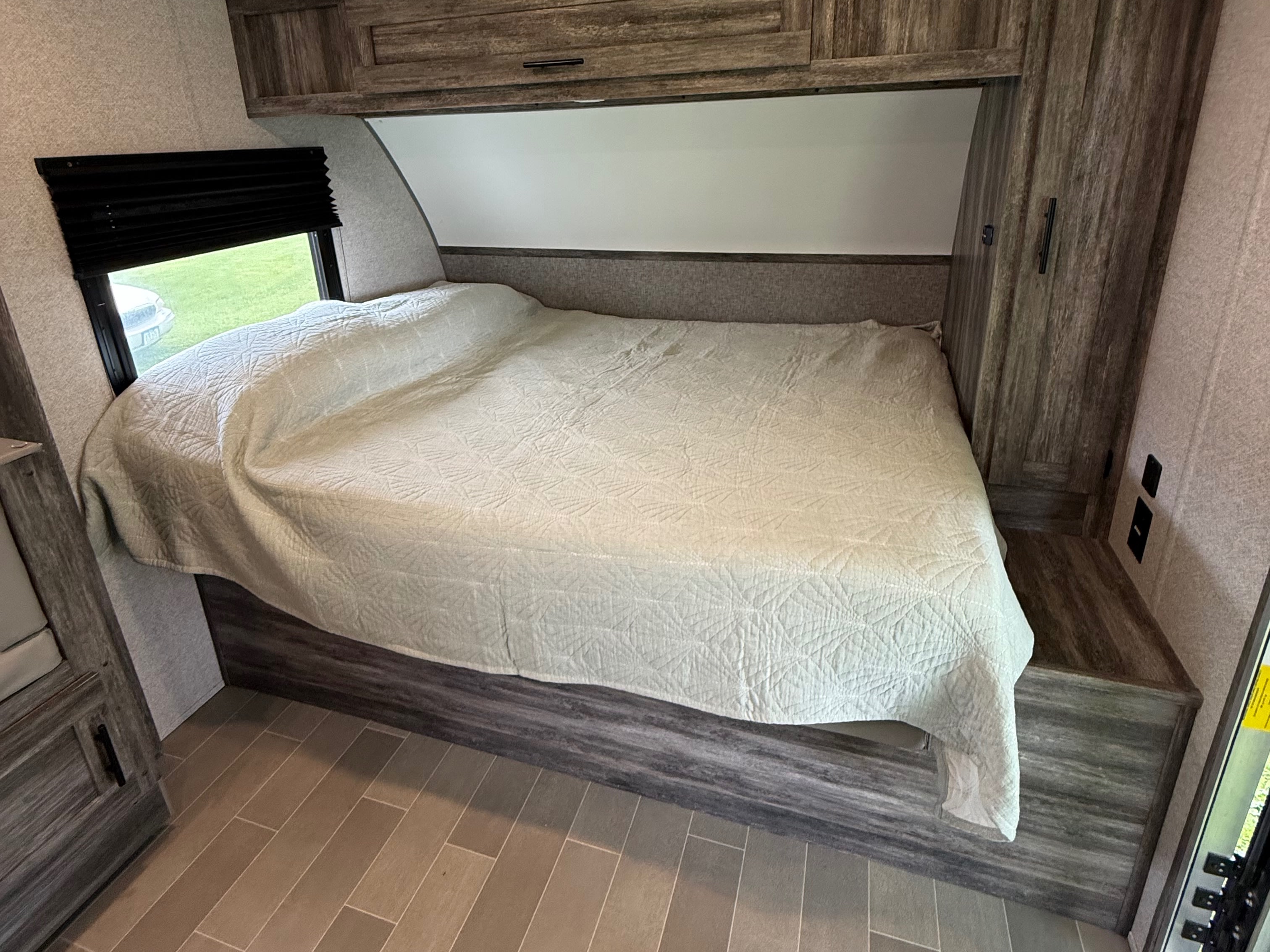 Cozy RV rental bedroom featuring a bed with light quilt, window with dark blind, wood cabinetry, and tiled floor.. Forest River Wildwood FSX Platinum 2021