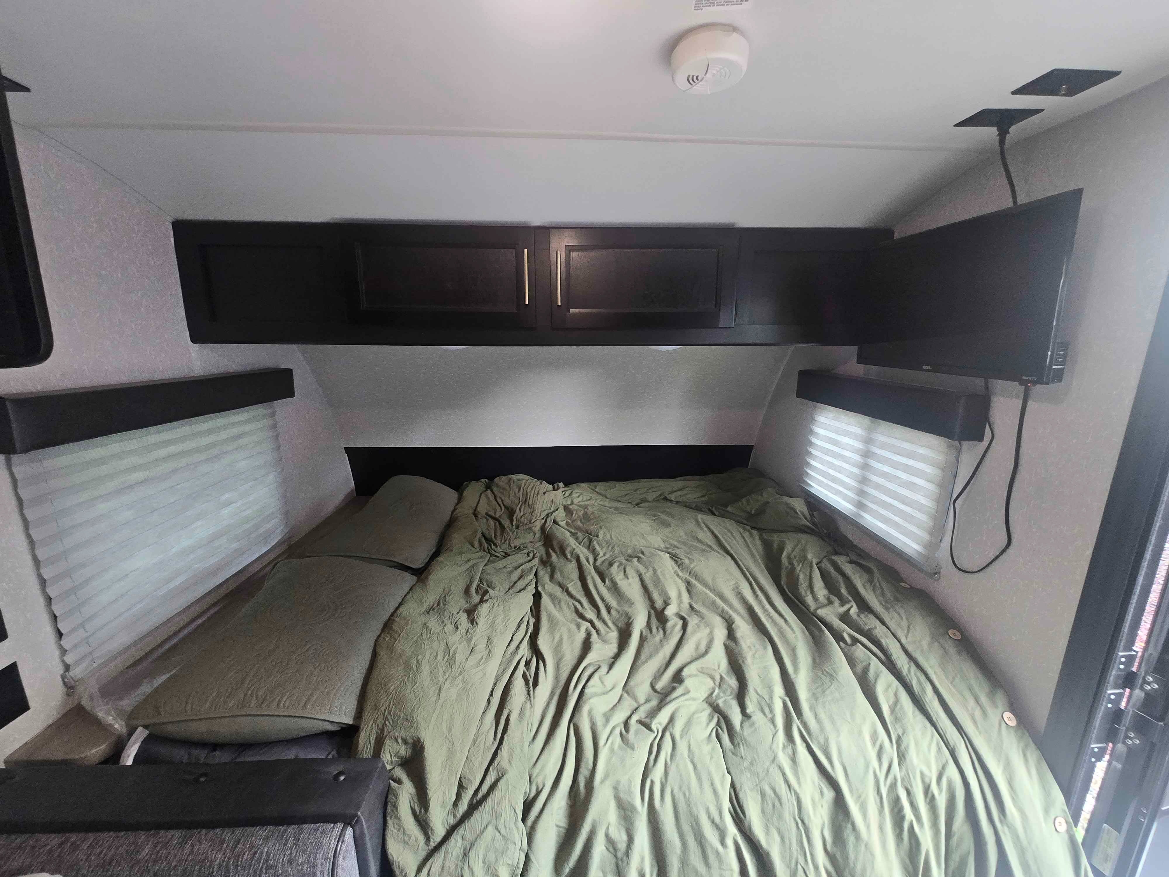 RV rental bedroom with a cozy bed, green comforter, overhead cabinets, wall-mounted TV, window blinds, and smoke detector.. Forest River Cherokee Wolf Pup 2022