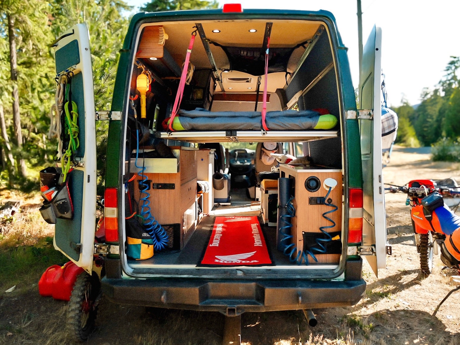 This RV rental camper van showcases its custom interior, featuring a bed, storage, and gear, with a dirt bike nearby.. Mercedes-Benz Sprinter 2006