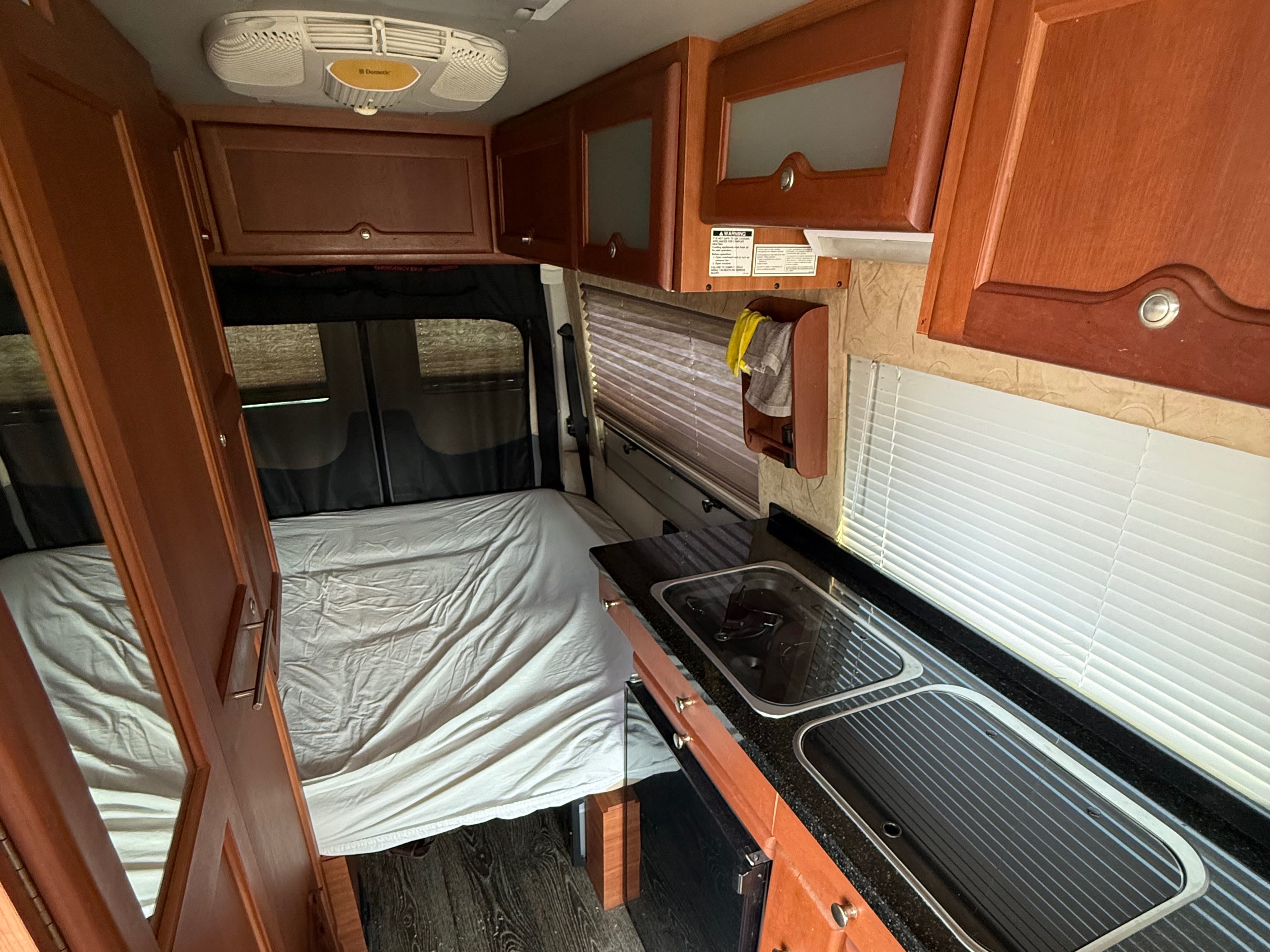 Cozy rv rental interior: kitchen with sink & bed in a compact camper van. Wooden cabinets and window blinds visible.. Roadtrek Roadtrek Class B 2014
