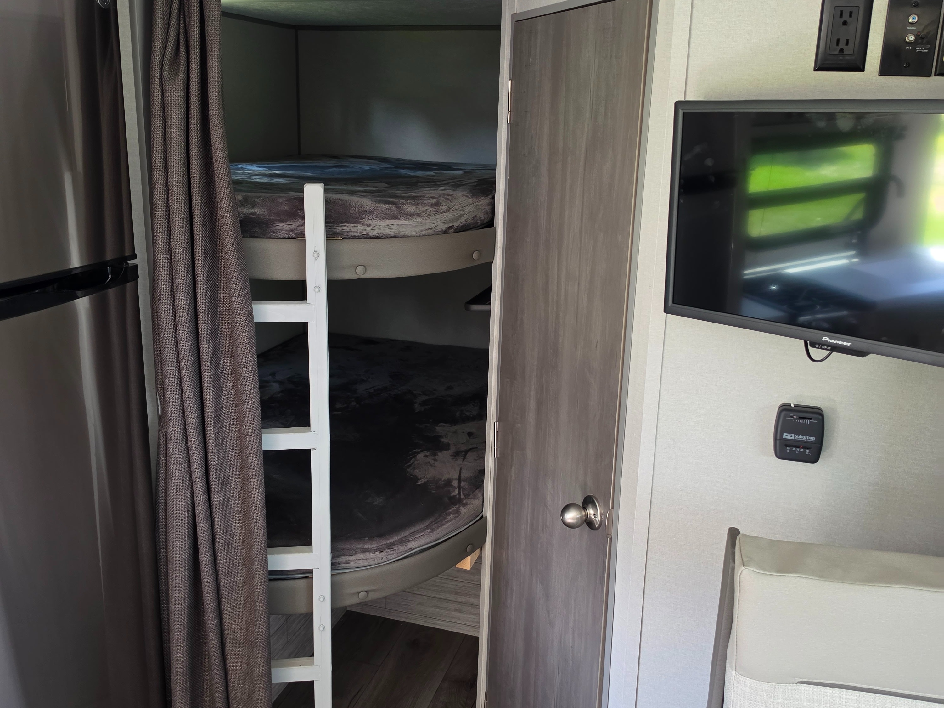 RV rental interior shows bunk beds with a ladder, a dark curtain, stainless steel fridge, TV, and wall thermostat.. Coachmen 184BHS 2024