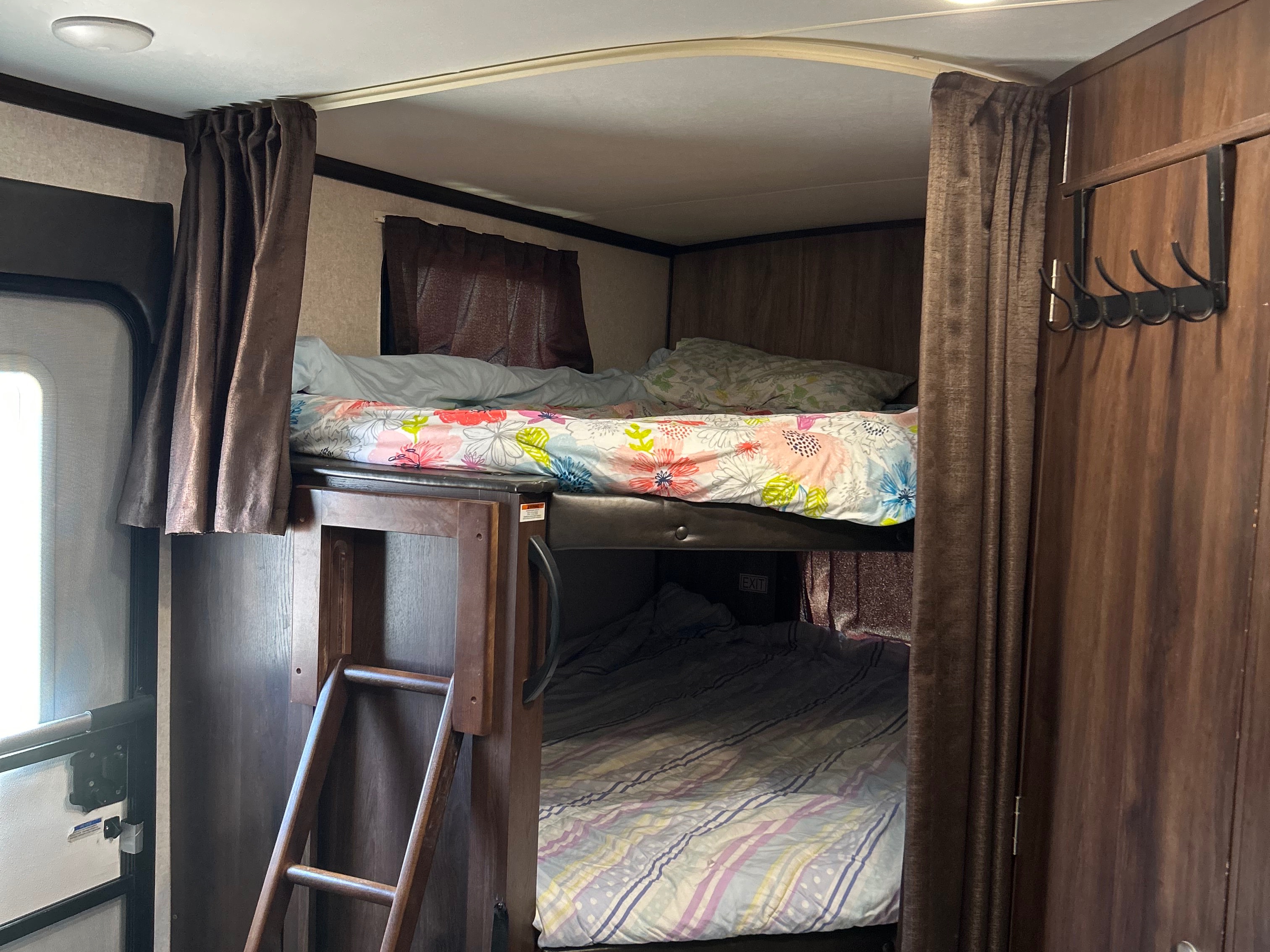 Inside an RV rental: two bunk beds with a ladder, floral & striped bedding, privacy curtains, & a coat rack.. Jayco White Hawk 2019