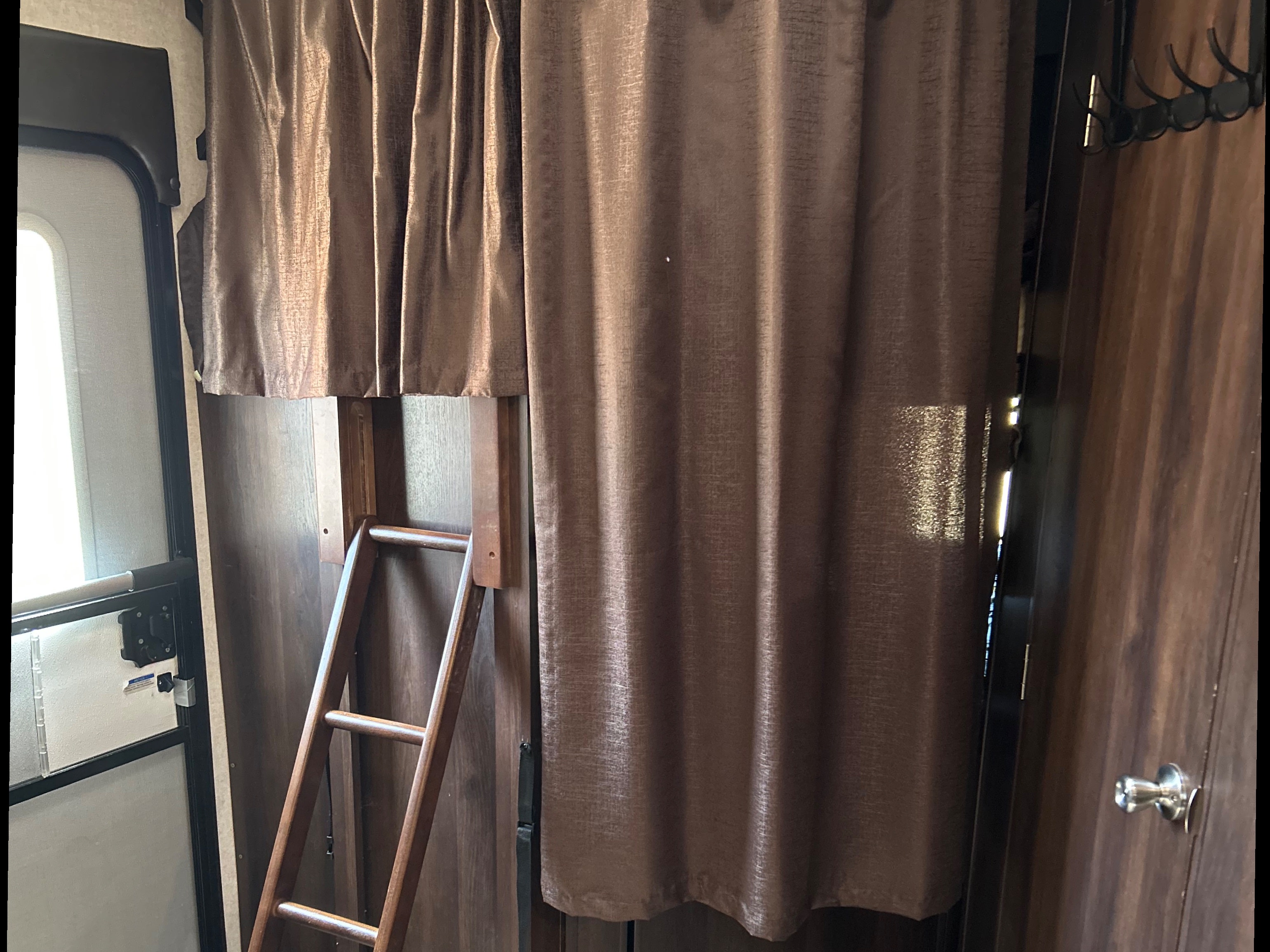 RV rental interior with a wooden bunk bed ladder, brown privacy curtains, a door with a silver knob, and a coat rack.. Jayco White Hawk 2019