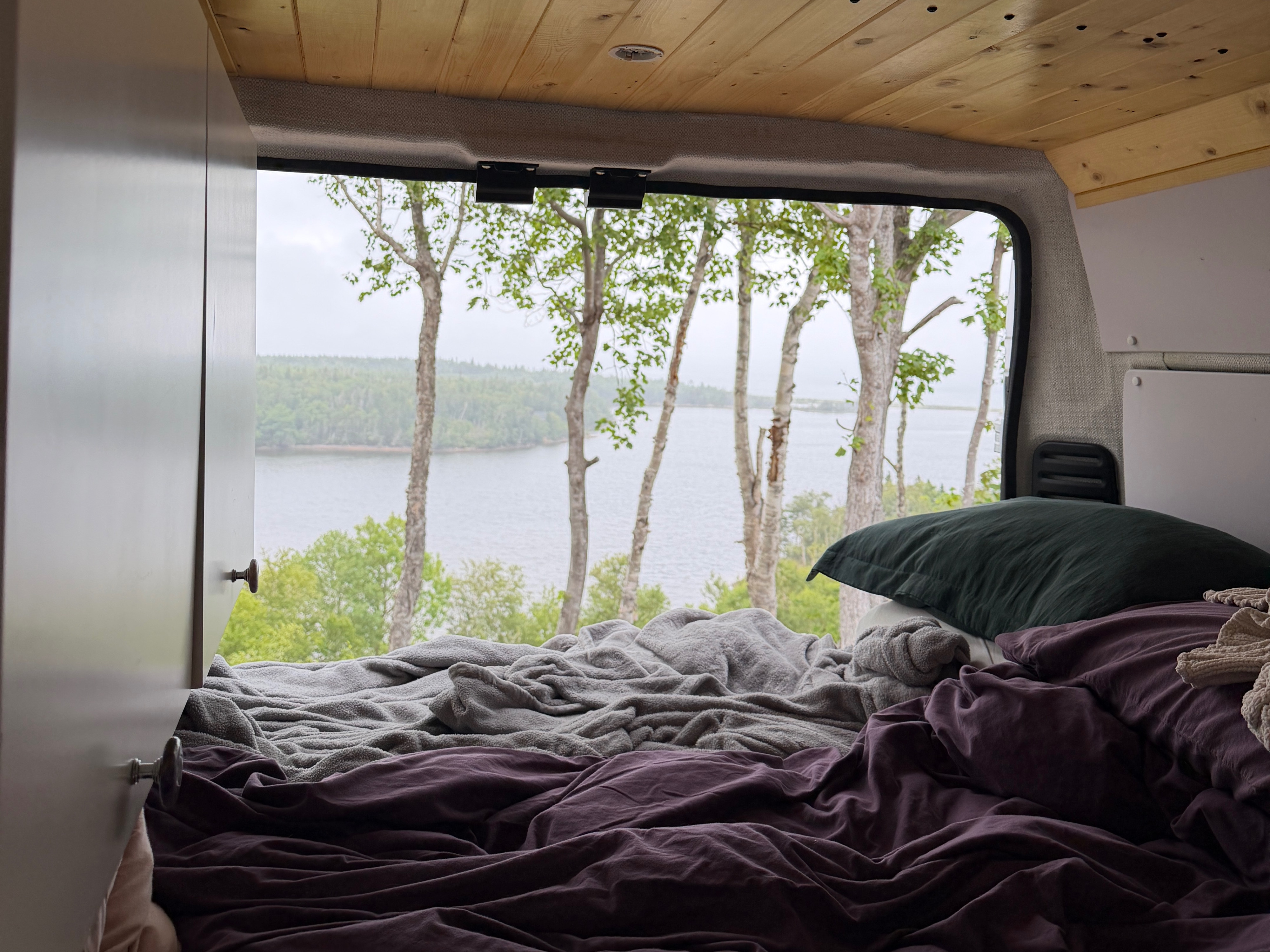 Cozy RV rental bedroom with a rumpled bed, dark green pillow, and a large window offering serene lake & forest views.. Ram Promaster 2024
