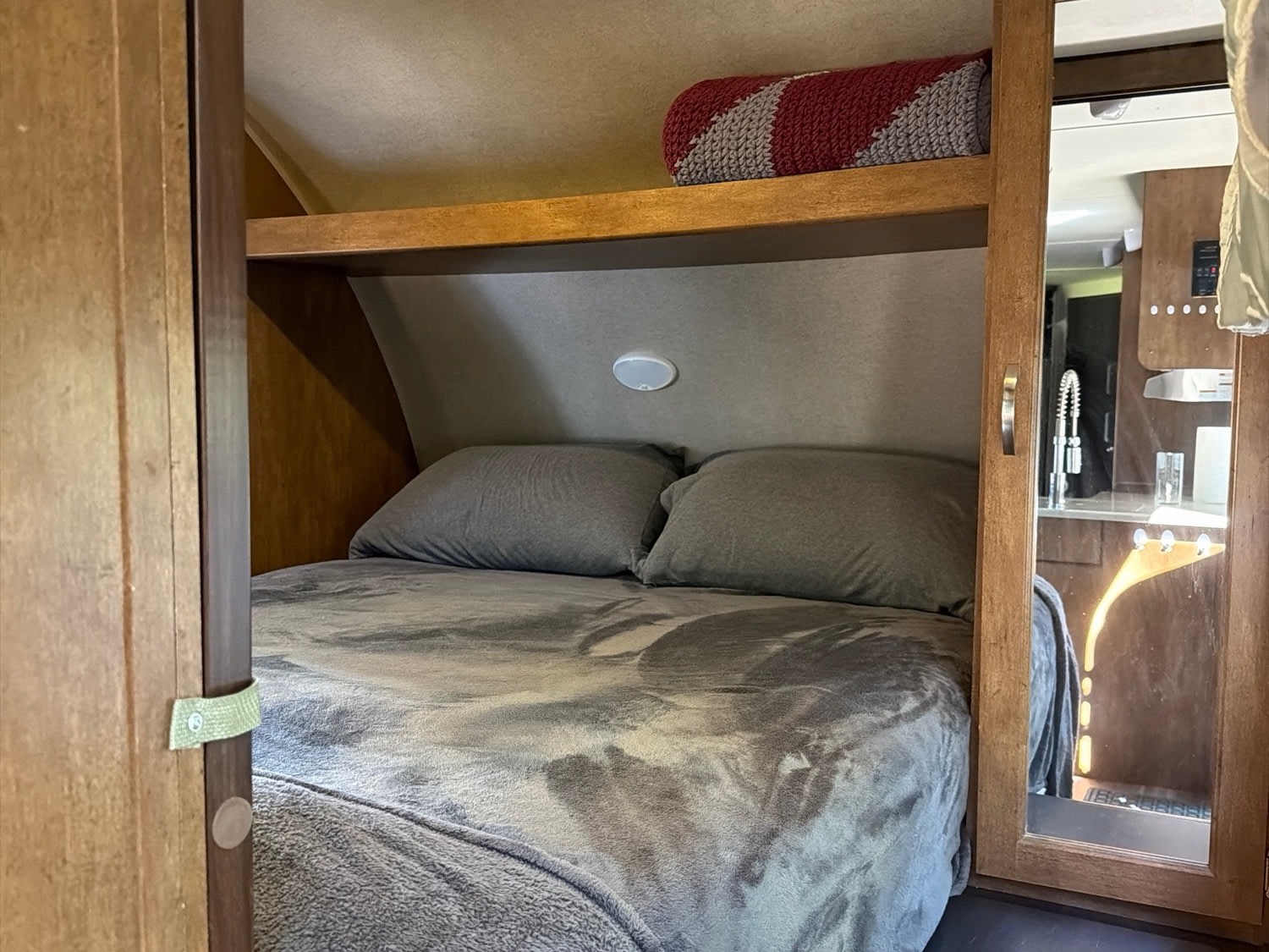 Compact RV rental bedroom featuring a cozy bed with gray bedding, pillows, and an overhead shelf. Mirror reflects the kitchen.. Forest River T27TDSS 2017