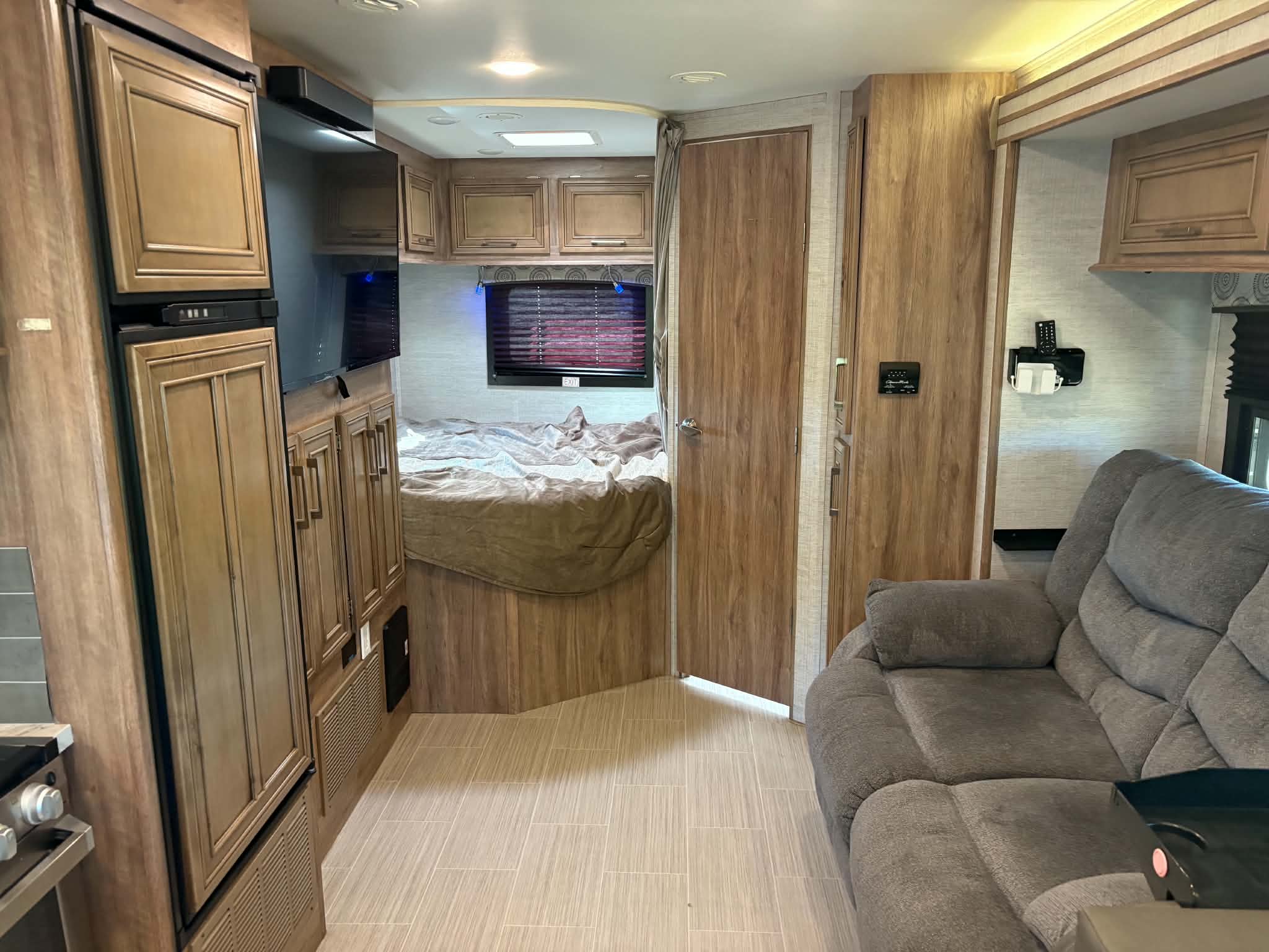 RV rental interior: A comfy bedroom, gray sofa, TV, fridge, & stove in a modern wood-paneled space. Ready for your next journey.. Entegra Coach Odyssey 2021