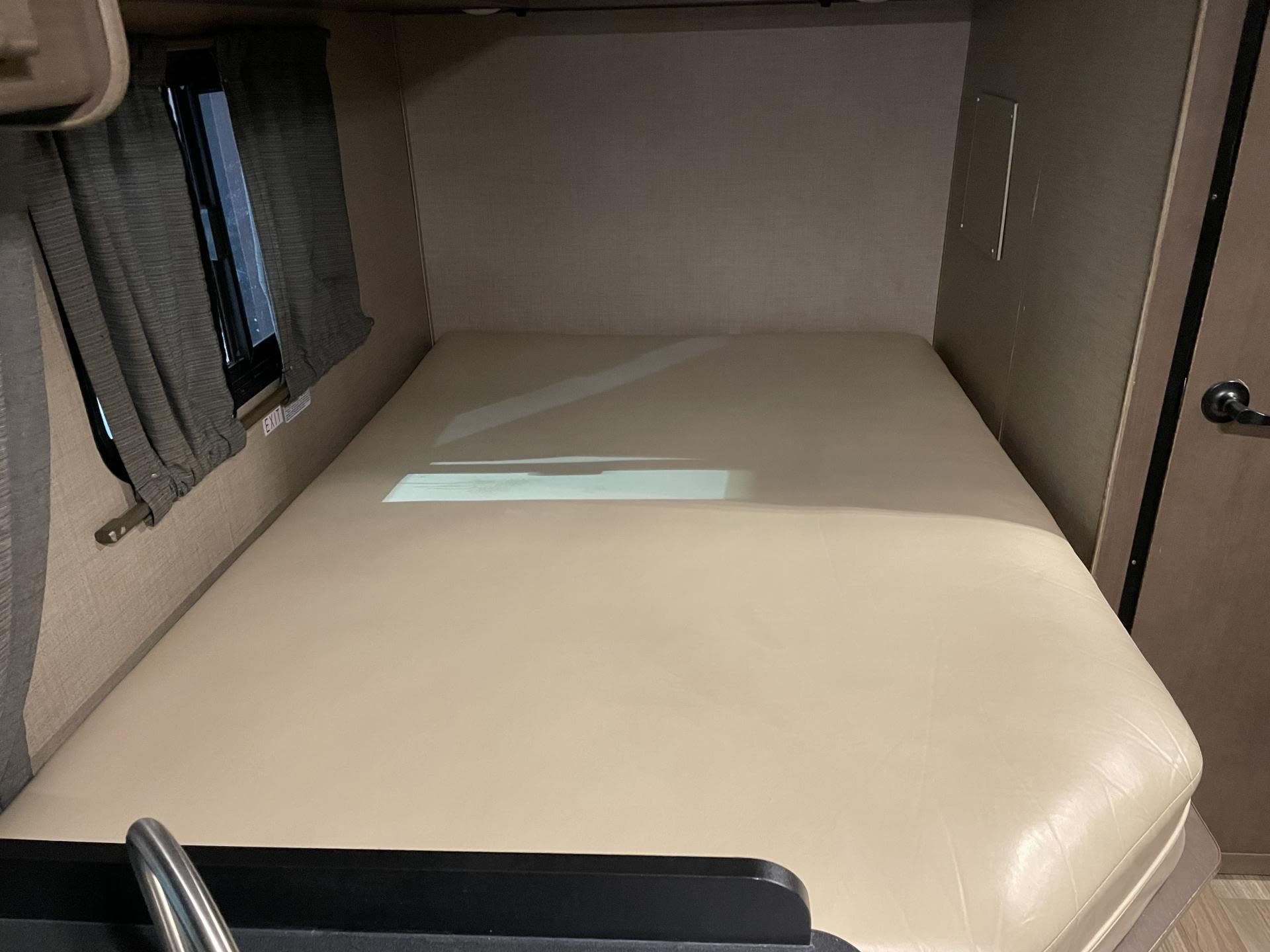 An RV rental bedroom with a bed featuring a cream-colored mattress, dark curtains on the window, and a wooden door.. Thor Majestic 2019