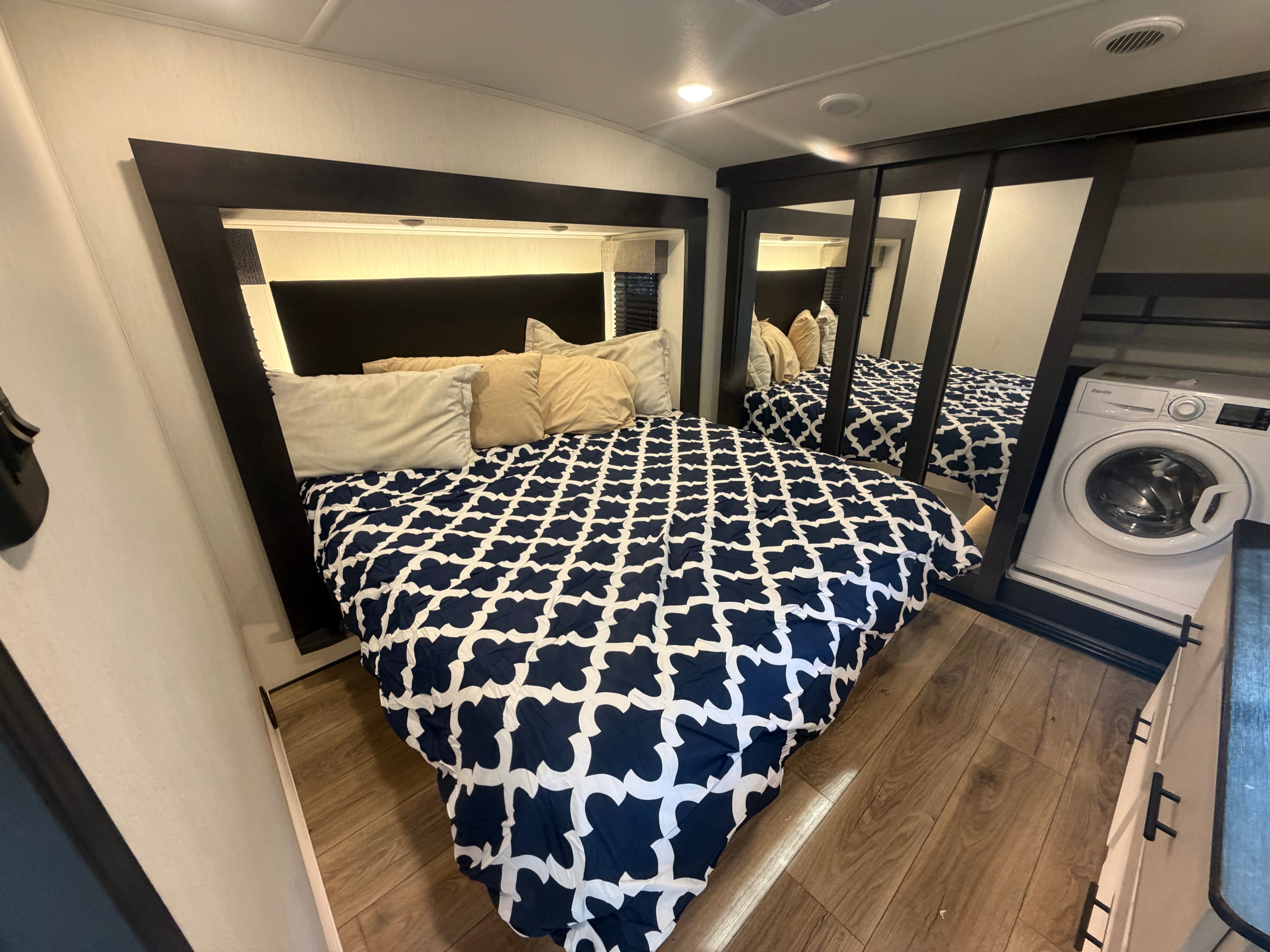 Cozy RV rental bedroom features a blue and white patterned bed, integrated lighting, a mirrored closet, and a compact washer.. Forest River Sabre 2025