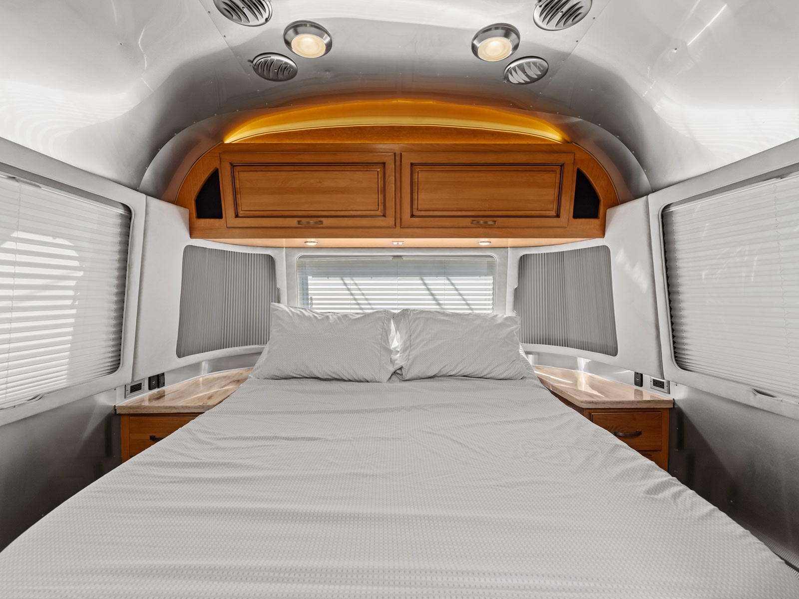 Cozy RV rental bedroom with a comfortable bed, overhead wooden cabinets, and windows with blinds in a metallic, modern interior.. Airstream Classic 2019