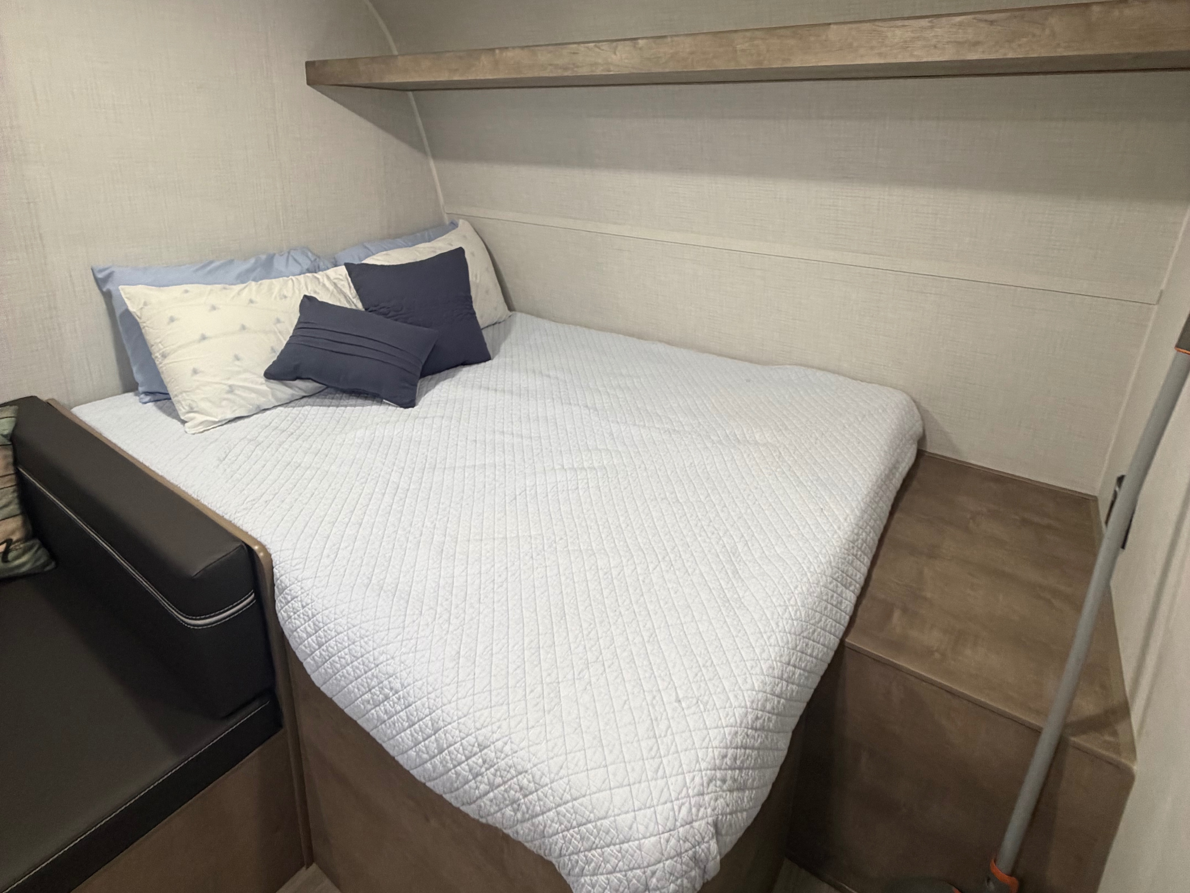 Inside an RV rental, a compact bedroom features a white quilted bed with blue and white pillows, an overhead shelf, and a dark bench.. Keystone RV Springdale Mini 2026