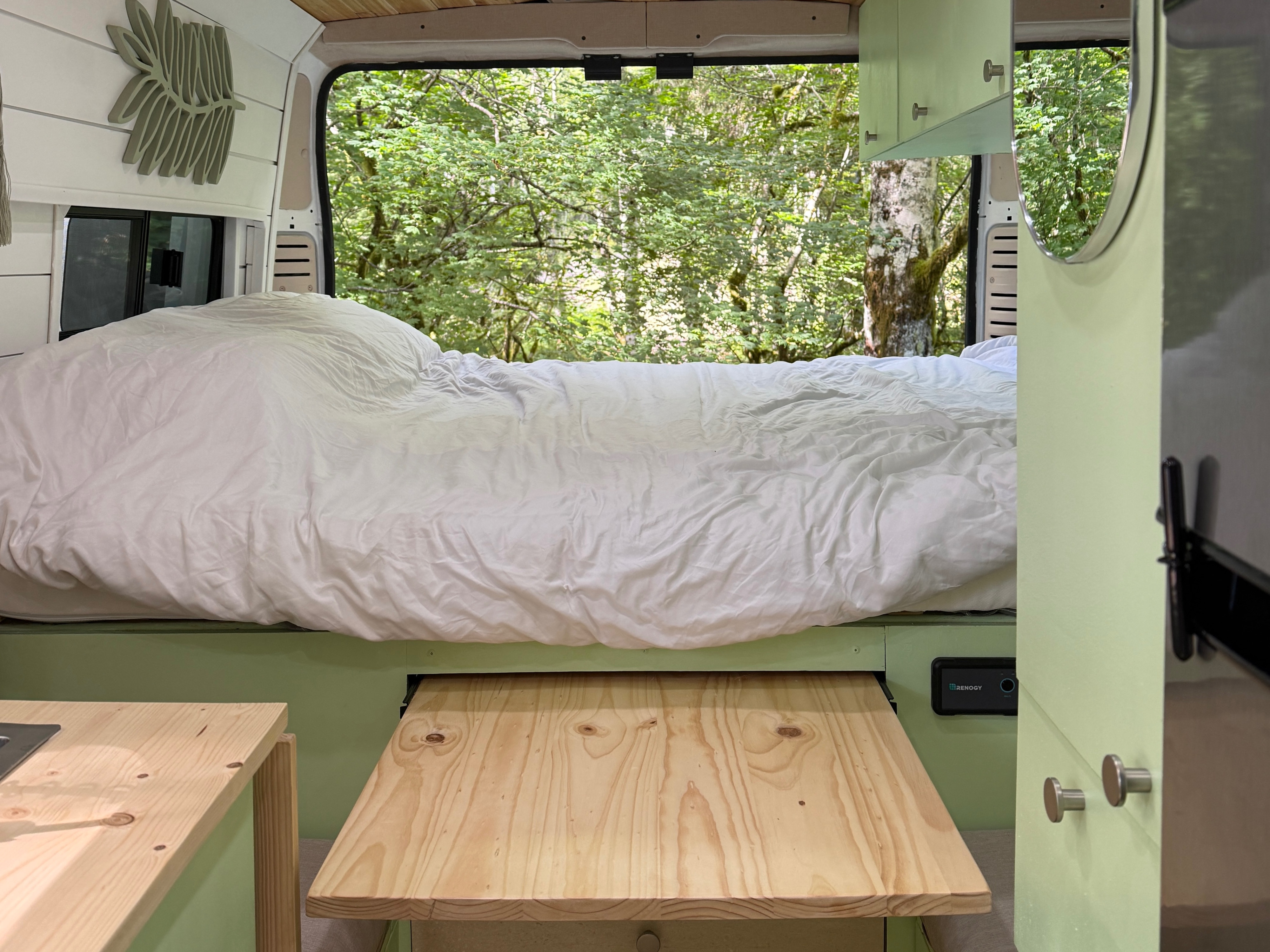 Cozy RV rental interior with a comfortable bed, pull-out table, and a scenic forest view out the back window.. Dodge Promaster 2023