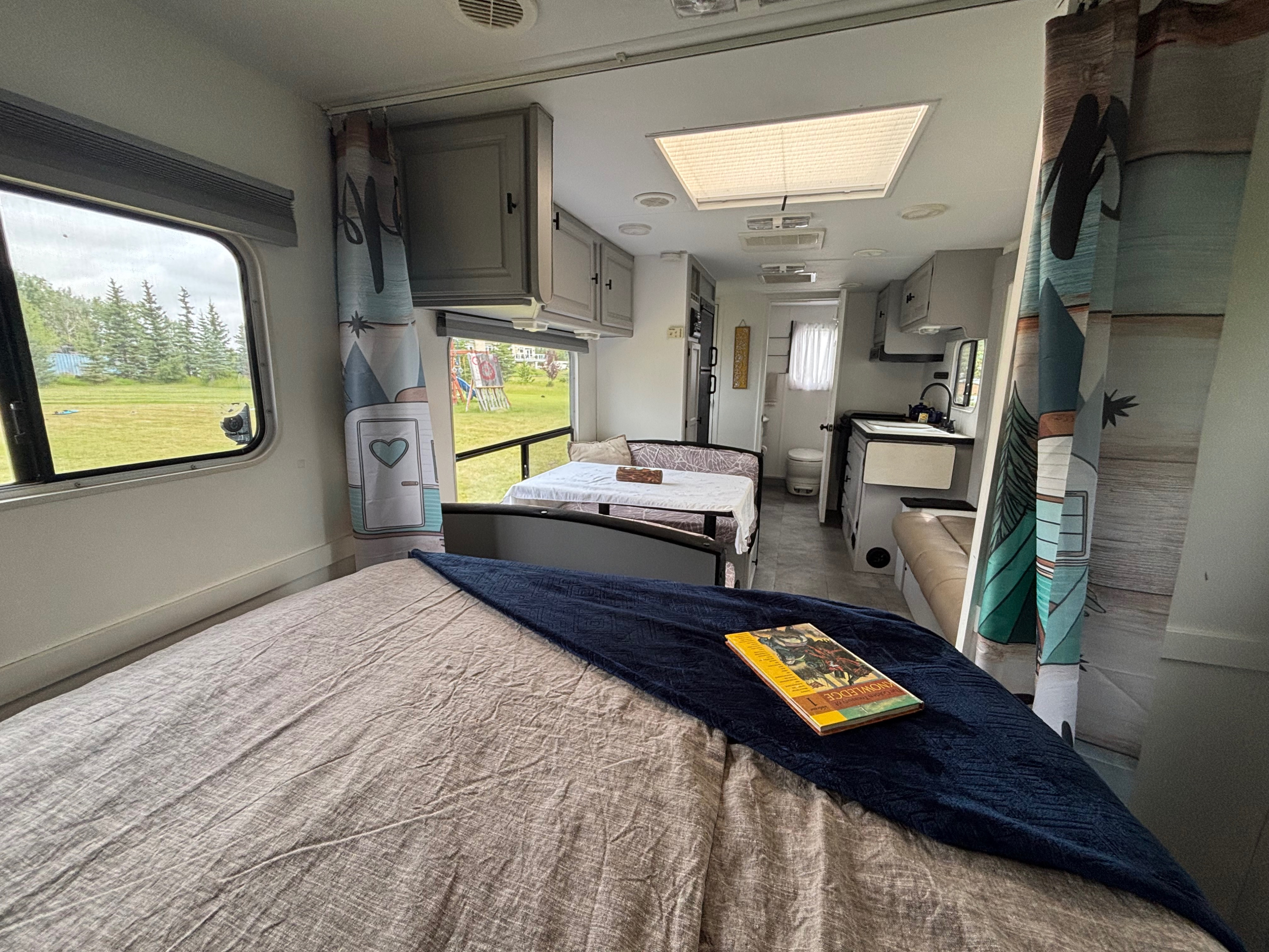 Cozy RV rental interior showing a bed, dinette, and kitchen area, with a lush green view outside the window. A book rests on the bed.. Kustom Koach Kustom Koach Trailer 2002