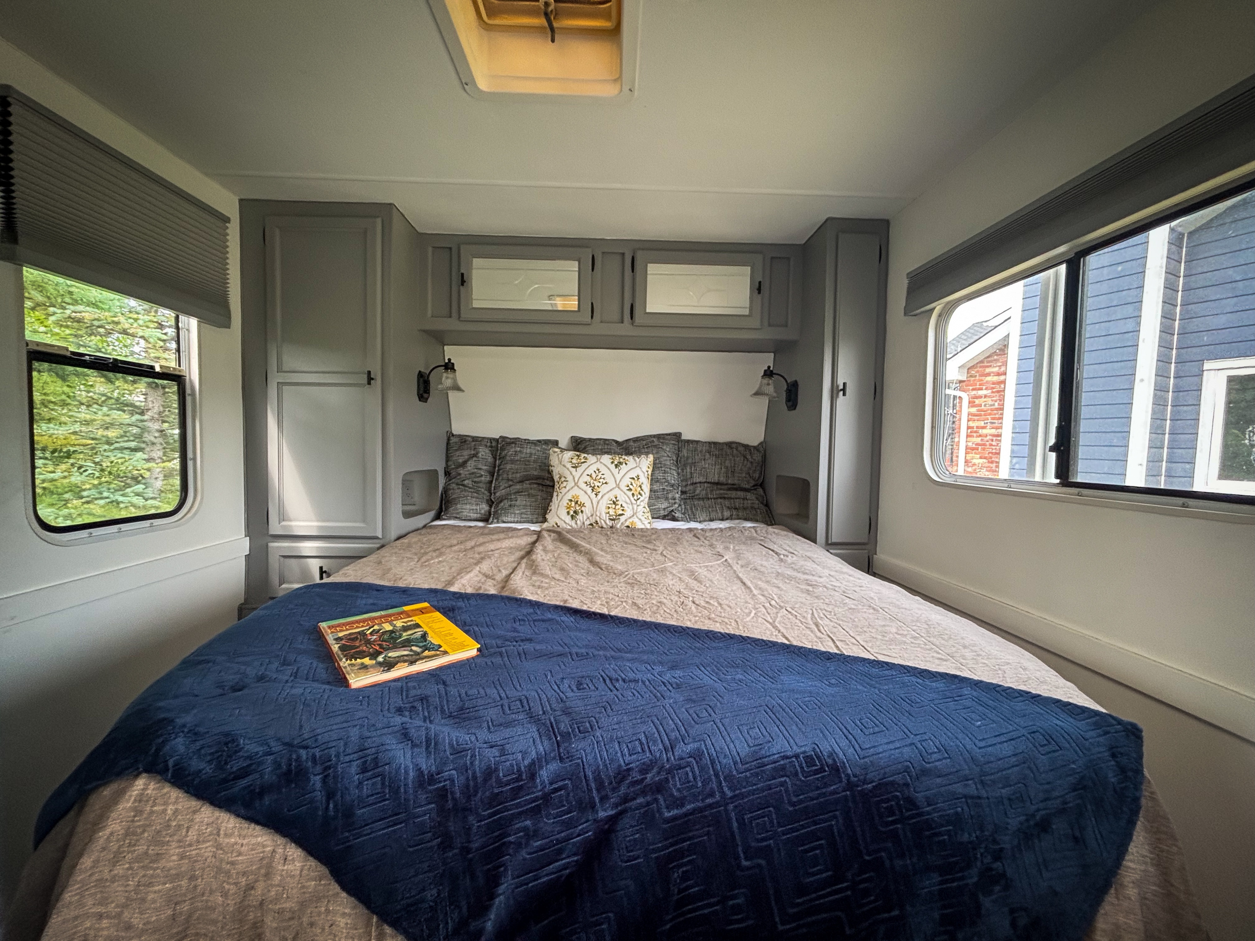 Cozy RV rental bedroom with gray cabinets, a comfortable bed with a blue blanket & book, and bright windows.. Kustom Koach Kustom Koach Trailer 2002