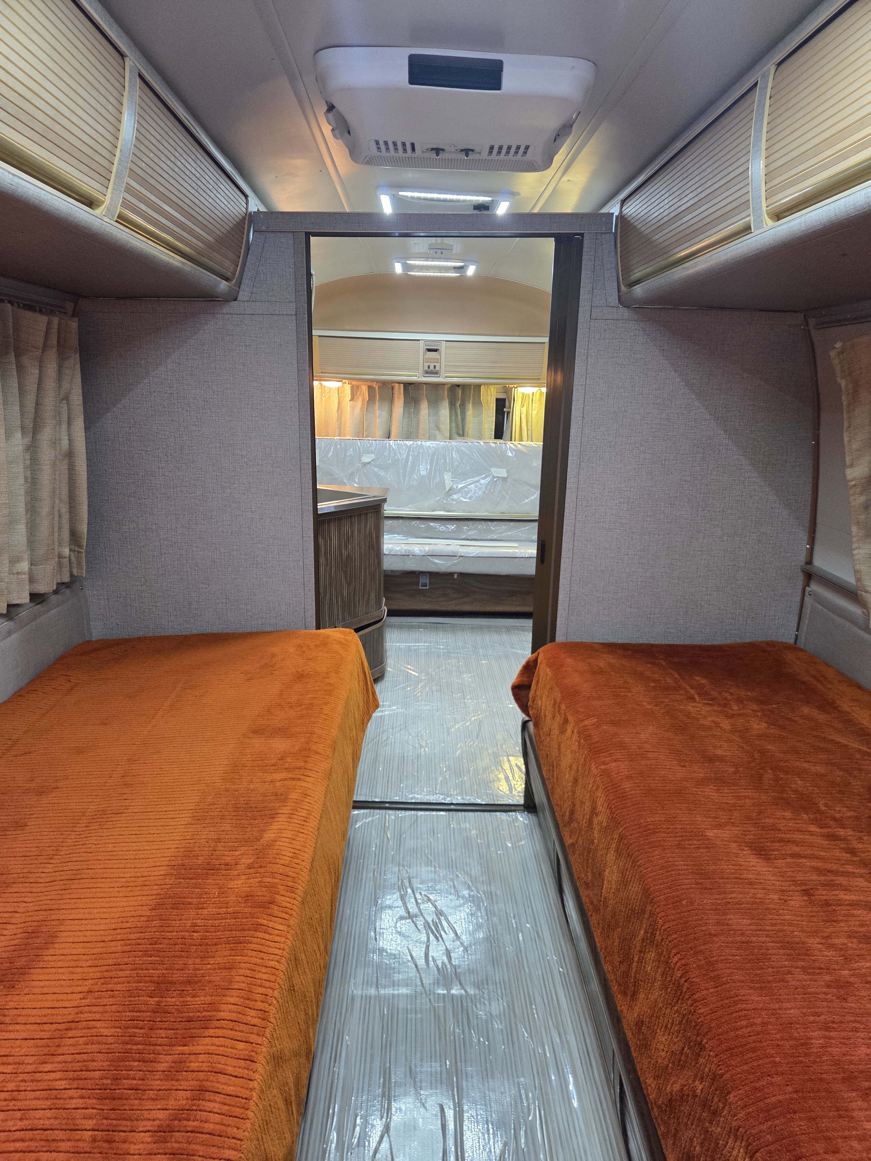Inside an RV rental: twin beds with bright orange blankets, pristine floor under protective film, and another sleeping area beyond.. Airstream Airstream Trailer 1977