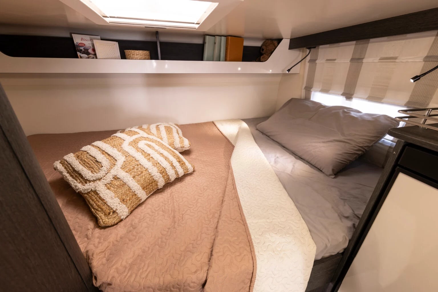 This rv rental bedroom offers a cozy bed with decorative pillows, built-in storage, and reading lights for your comfort on the road.. FORD TRANSIT 2026