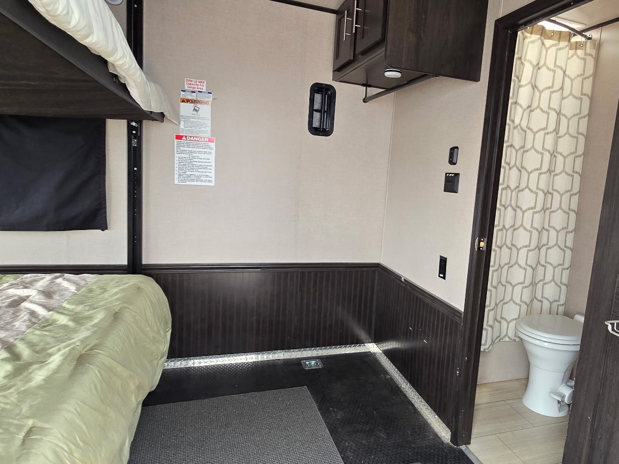 RV rental interior: bunk bed with green comforter, upper cabinets, and a bathroom with a toilet and patterned shower.. Forest River XLR HyperLite Toy Hauler 2020