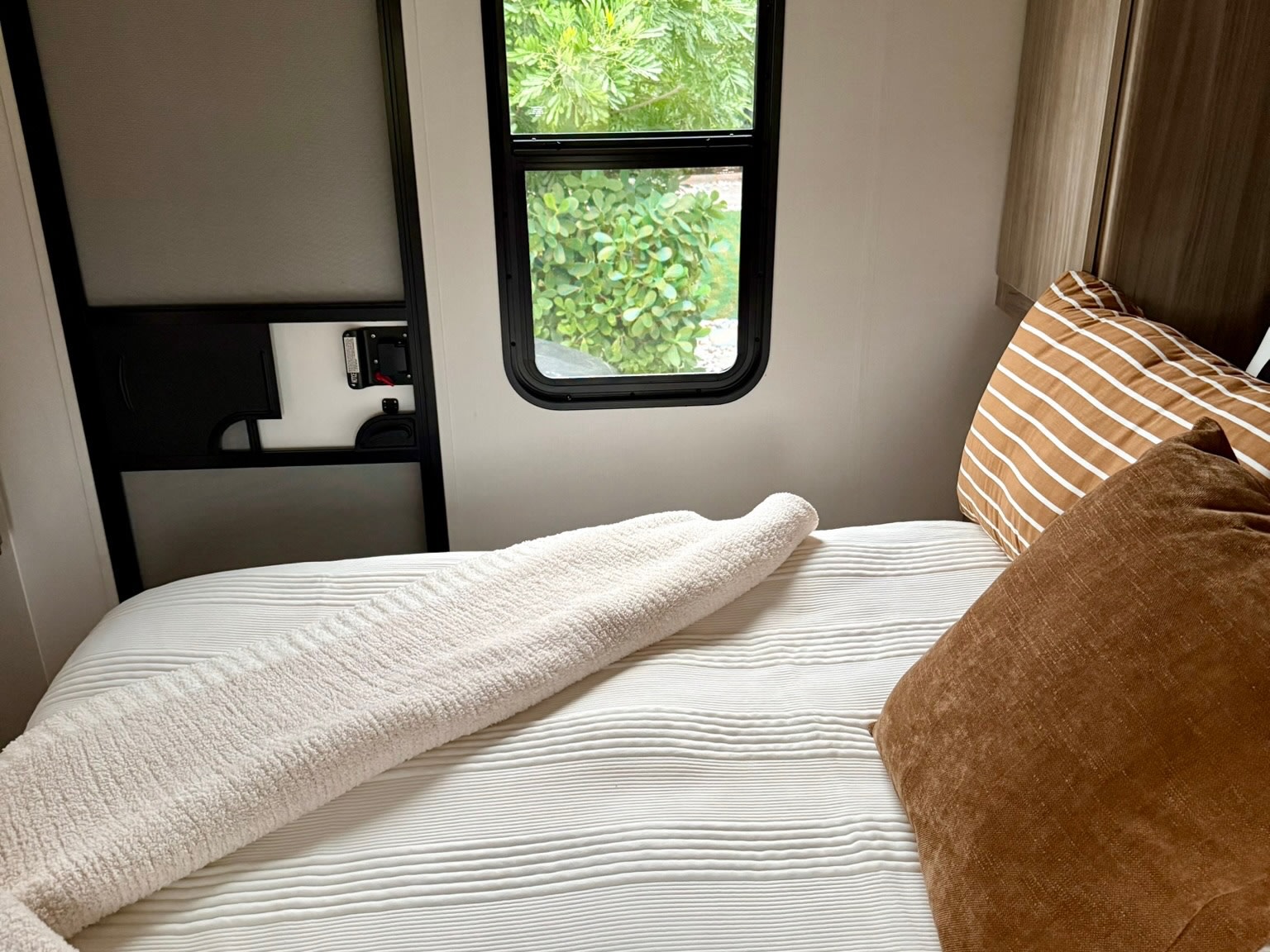 Cozy RV rental bedroom featuring a striped pillow, white textured bedding, and a window with a green nature view.. Winnebago Access 2024
