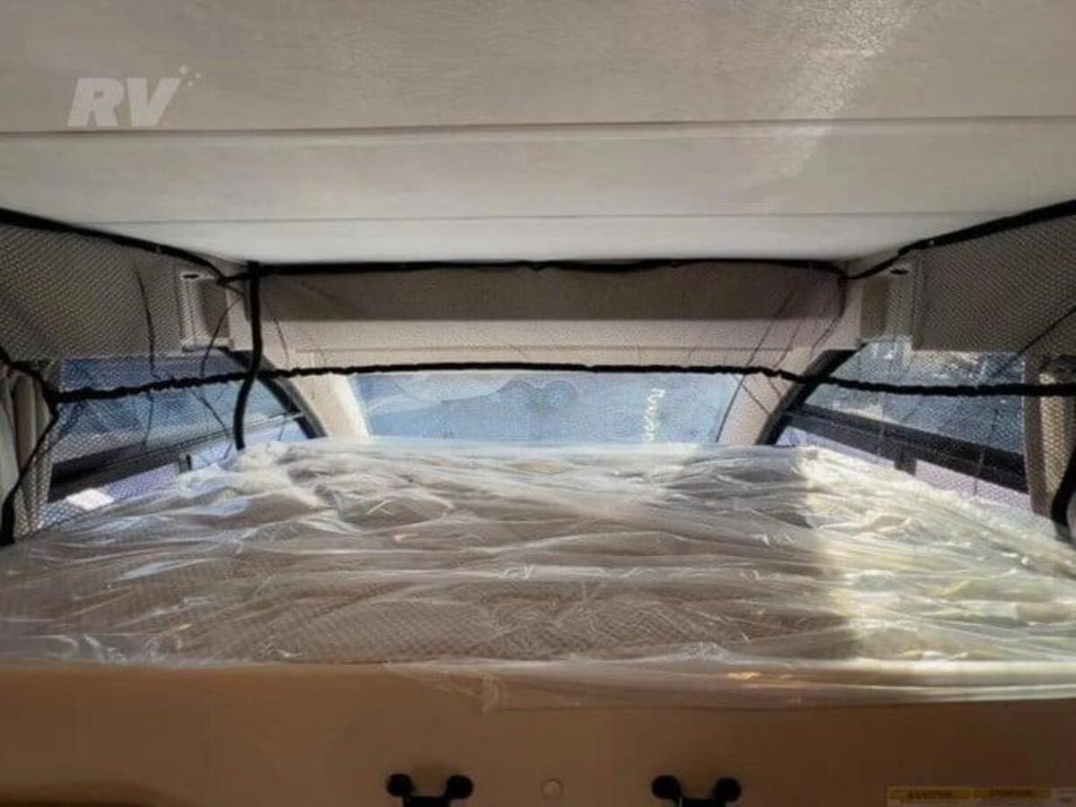 Inside an RV rental: a cab-over bed with a mattress still in its protective plastic wrap and a safety net above.. Thor Axis 2023