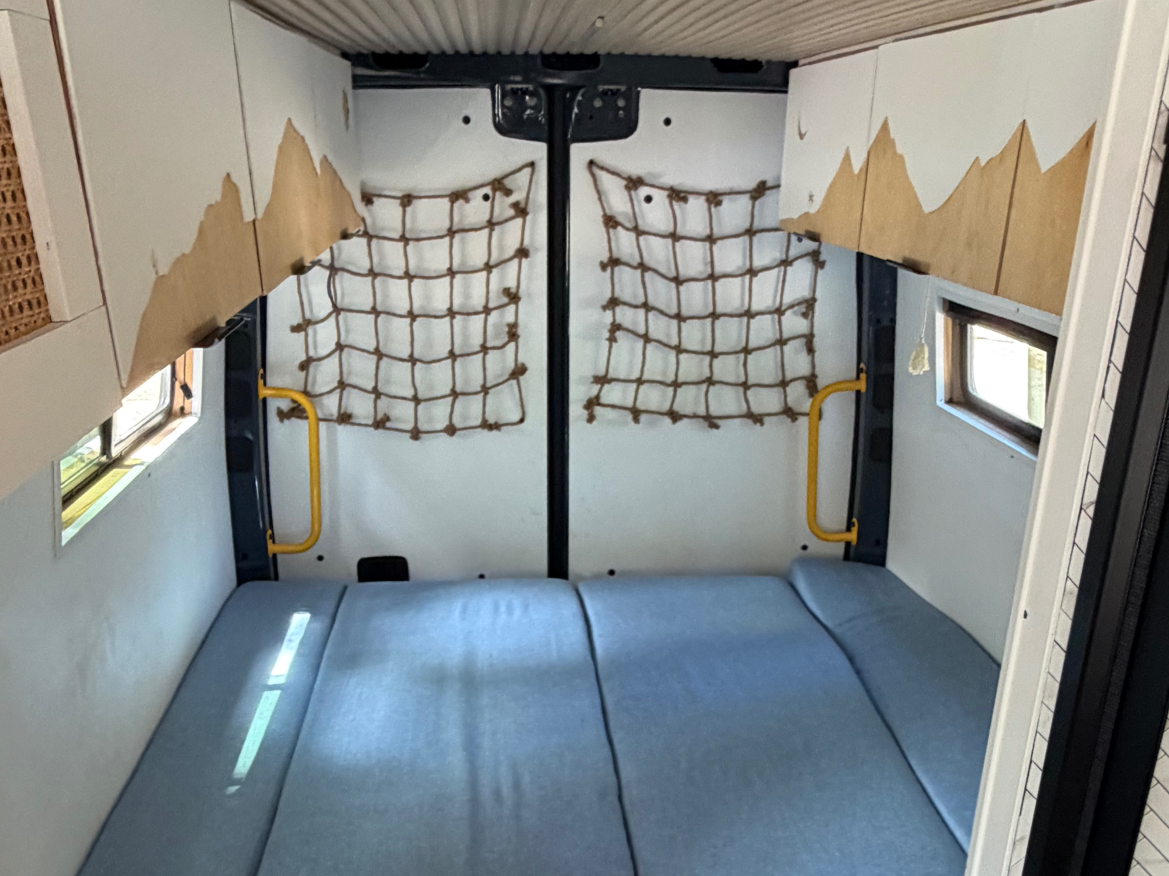 Cozy RV rental interior: blue mattress, small windows, cargo nets, and mountain-themed upper wall decor in the sleeping area.. Mercedes-Benz Sprinter 2019