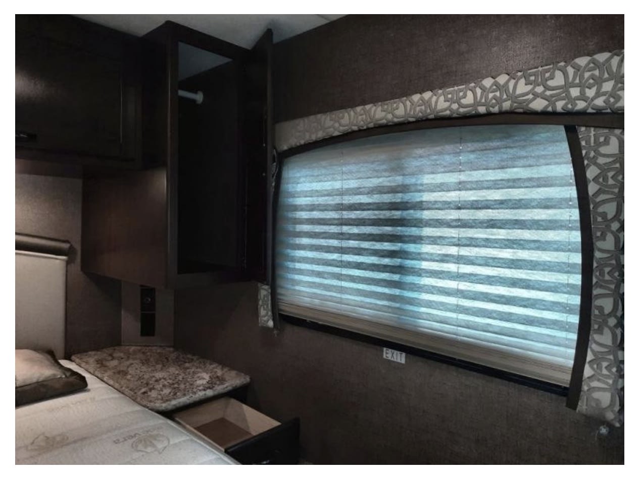 RV rental interior showing a bedroom area with dark wood cabinets, a bed, and a window with blinds and a decorative valance.. Thor Chateau 2020