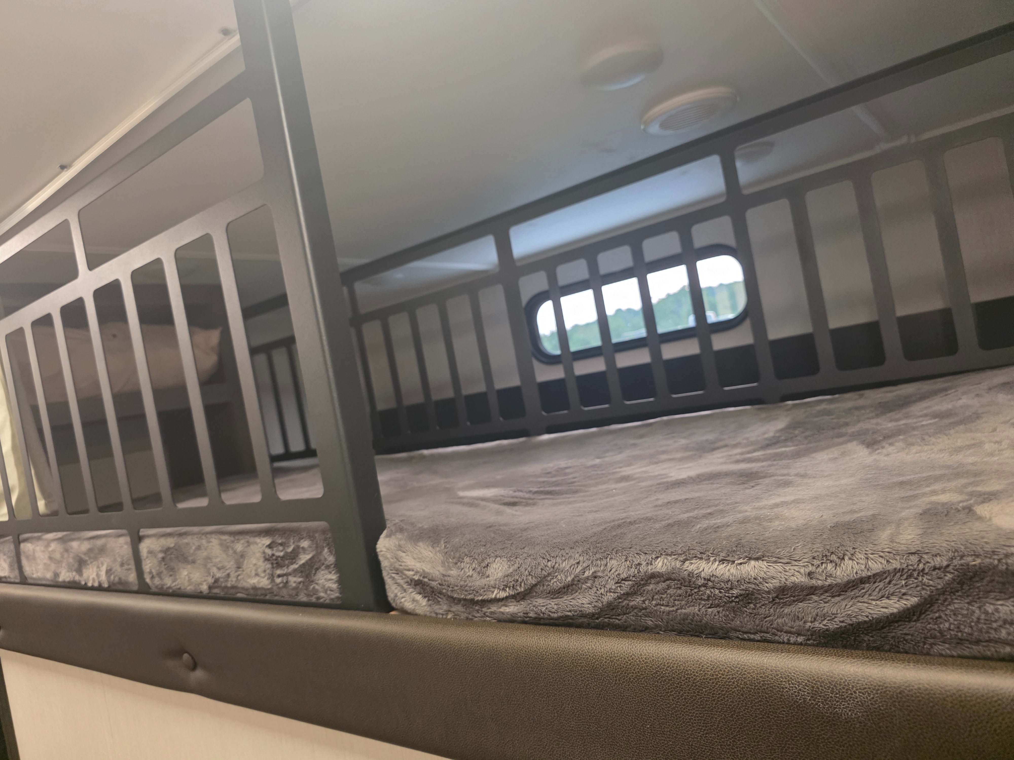 Inside an RV rental, showing compact bunk beds with soft gray blankets. An oval window reveals green scenery.. Forest River Cherokee Alpha Wolf 2026